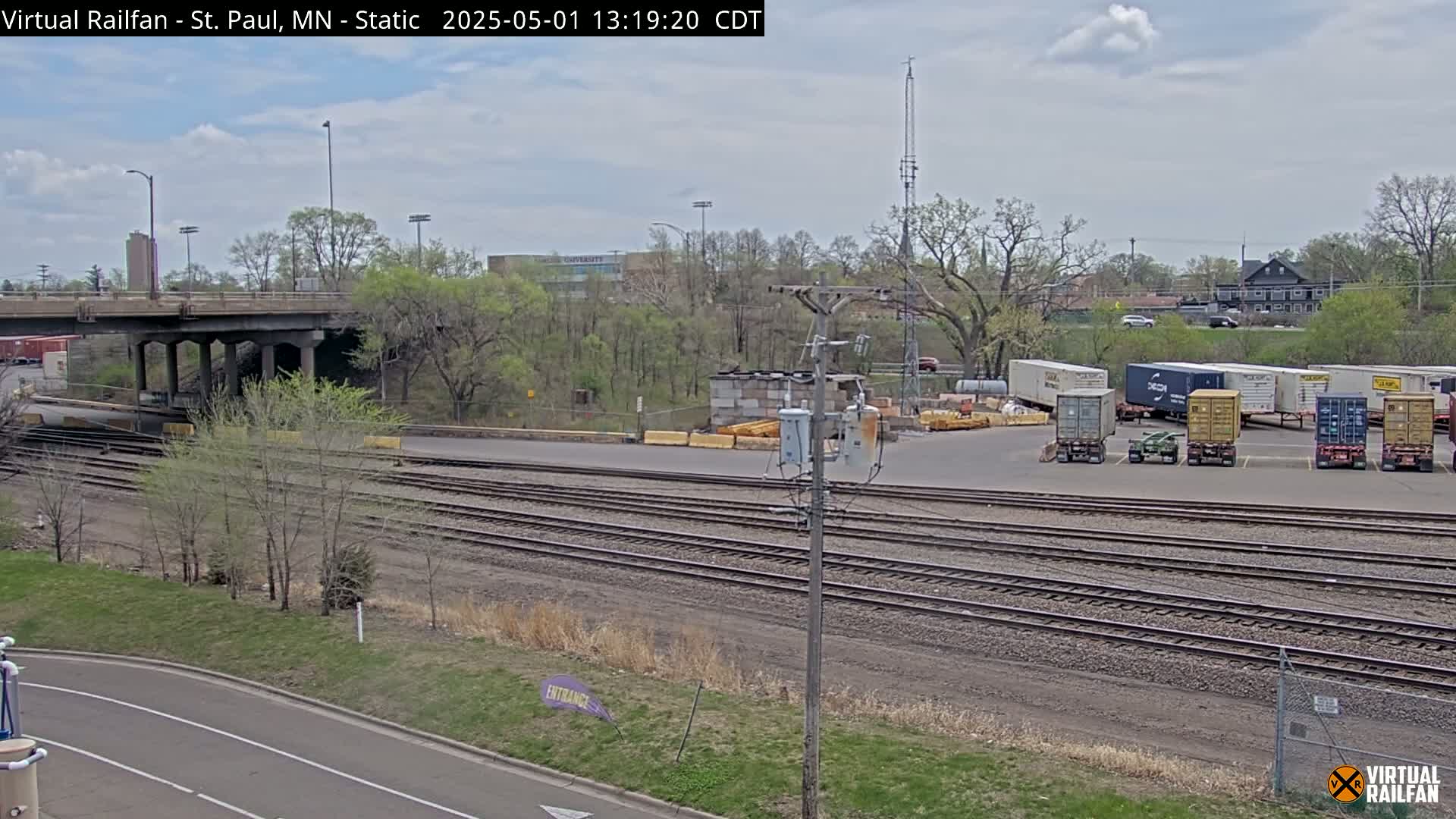 A partly cloudy day shows a view of railroad tracks, a bridge, and several shipping containers parked in a lot near a roadway and leafy trees.