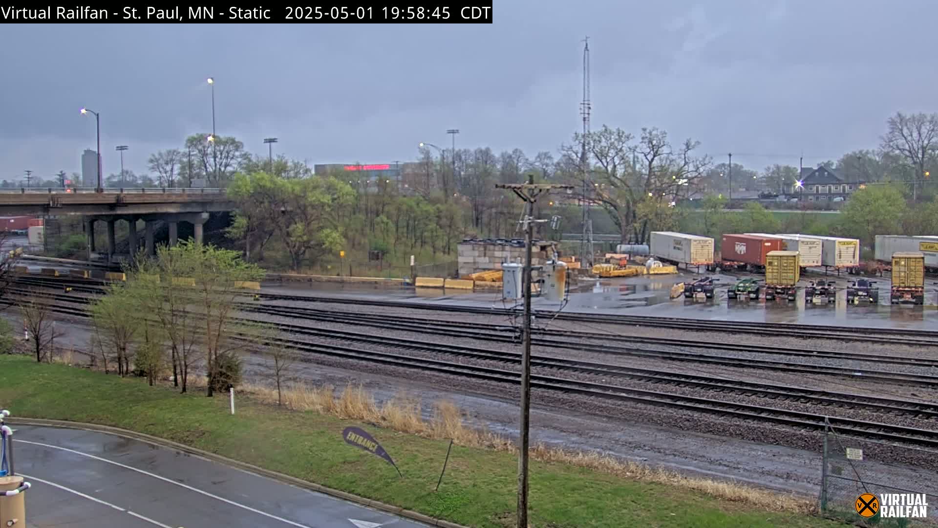 A wet, overcast scene shows a railway yard with multiple sets of train tracks, several shipping containers, and a highway overpass in the background.