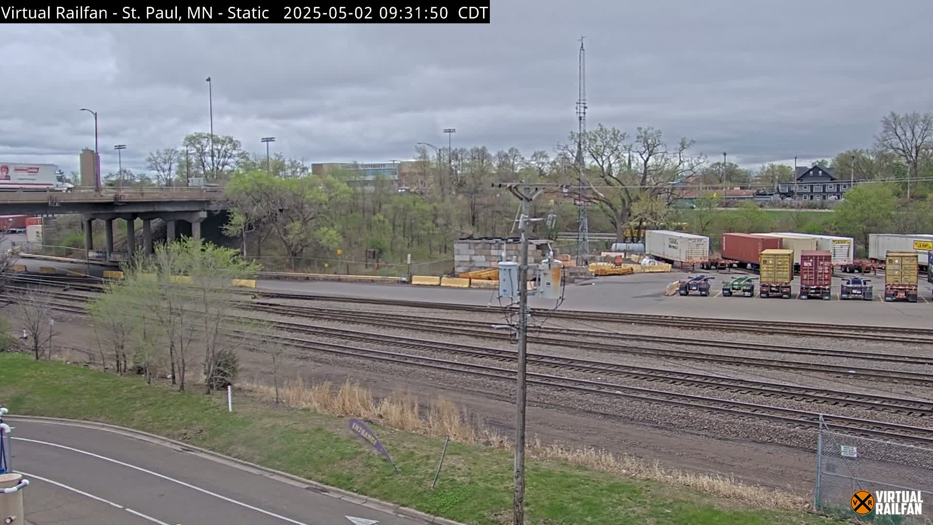 A mostly cloudy day reveals a railroad yard with several shipping containers and train tracks, a bridge overhead, and green vegetation in the background.