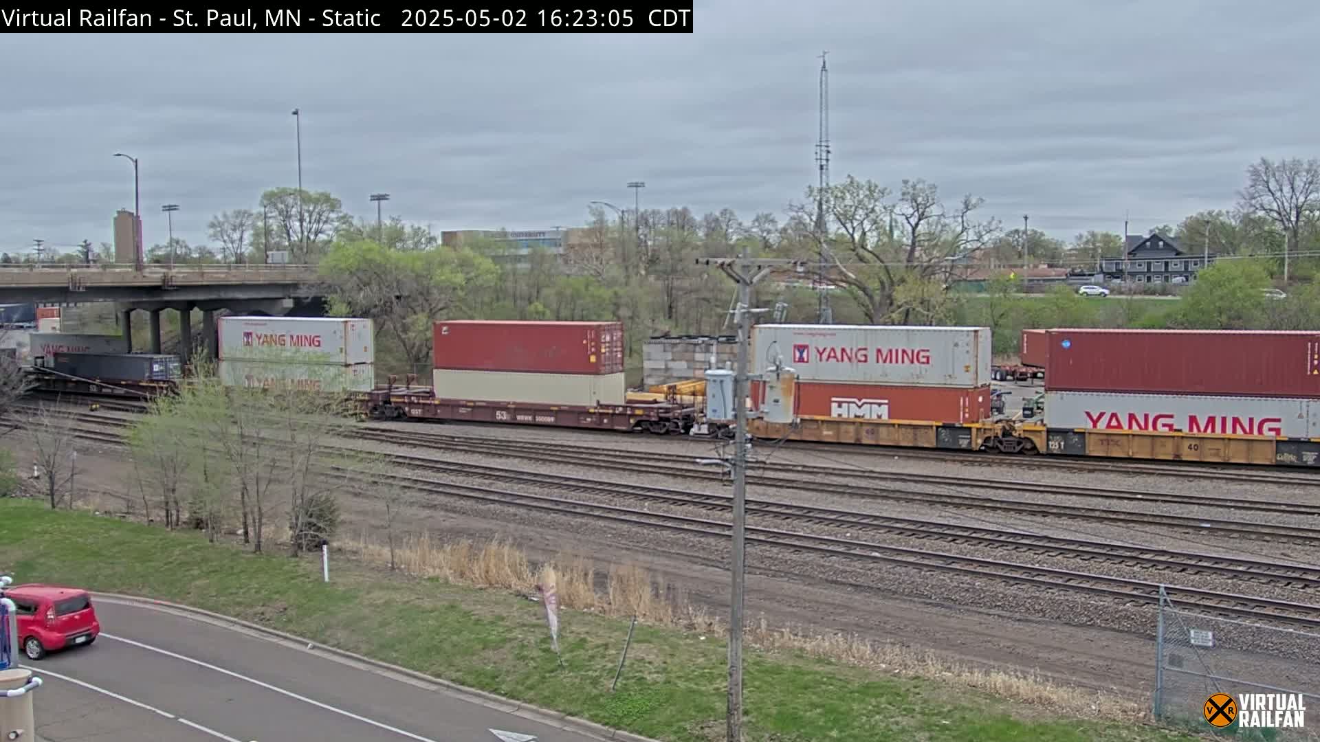 Under a cloudy sky, a freight train carrying multiple shipping containers labeled "Yang Ming" is visible on a railway track next to a road.