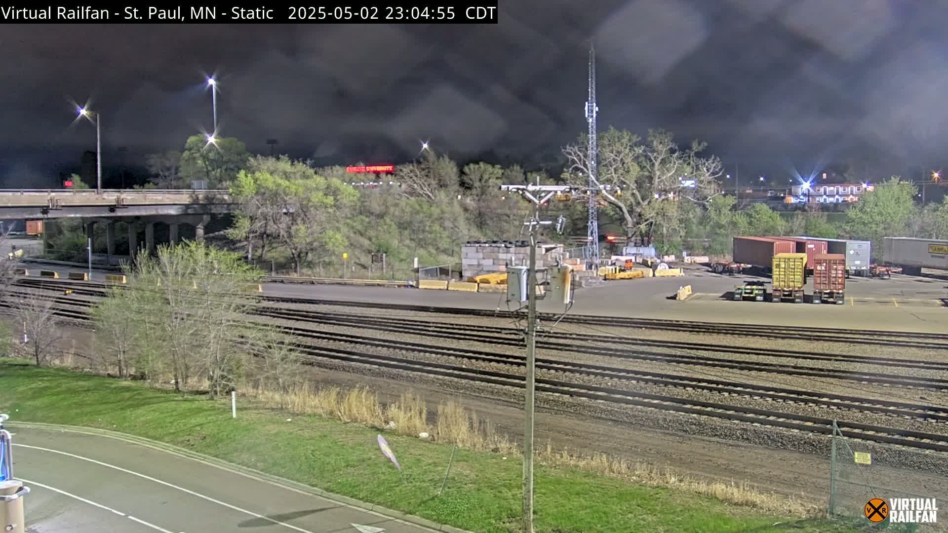 A nighttime view of railroad tracks, a roadway, and several shipping containers in a rail yard under a cloudy sky.