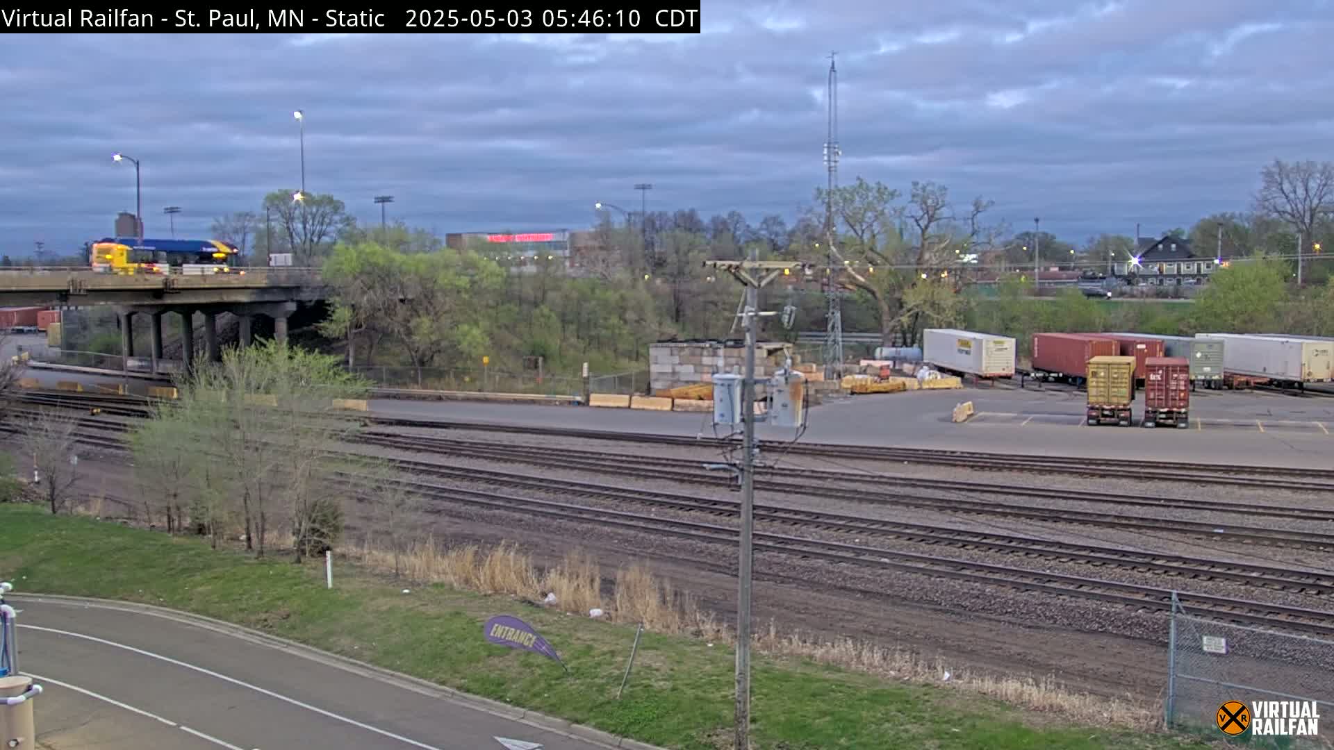 A bus drives across an overpass above railroad tracks and a switching yard under an overcast sky.