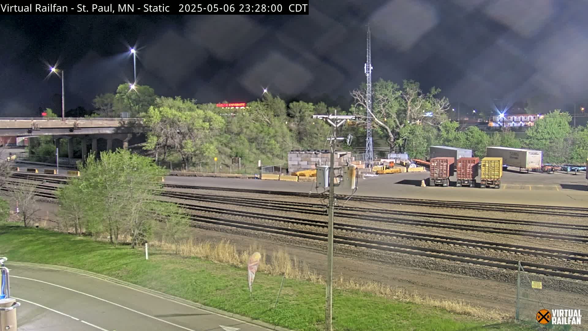 Under a dark, nighttime sky, a rail yard with several shipping containers sits beside a road and train tracks, bordered by trees and greenery.
