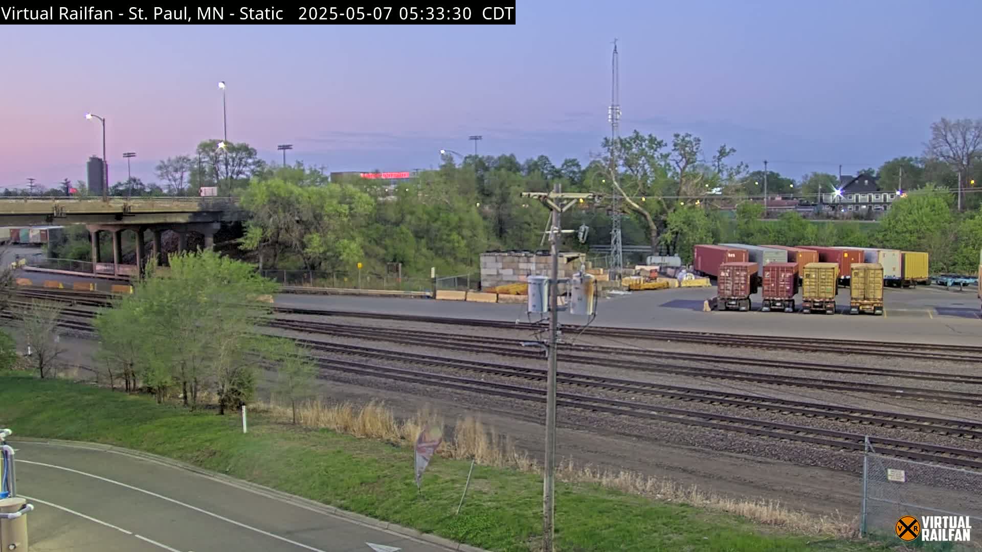 A twilight view of railroad tracks and several shipping containers near a highway overpass, with trees and greenery surrounding the area.