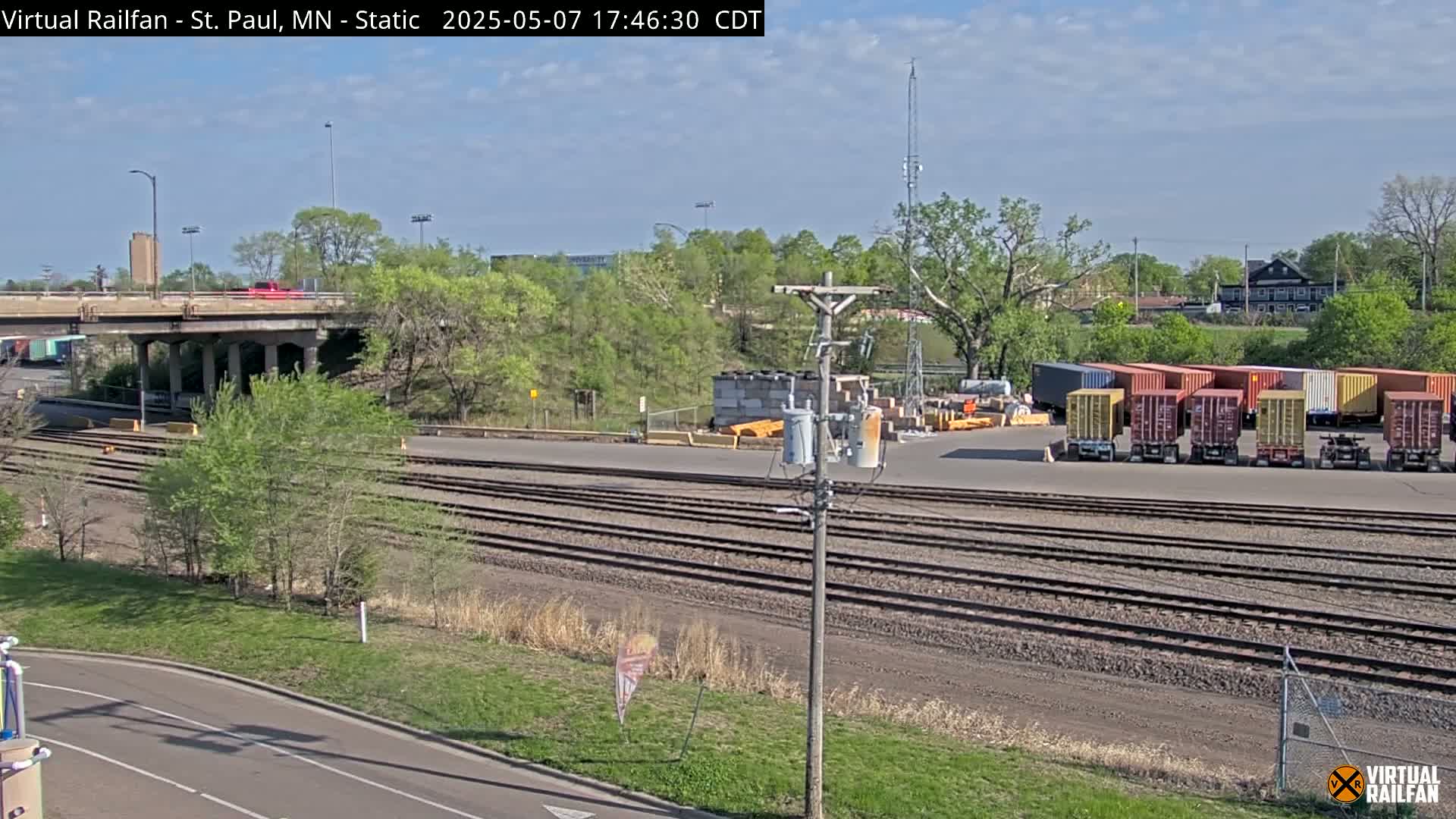 A sunny day reveals a railway yard with several shipping containers, a road with a car crossing a bridge overhead, and trees in leaf.