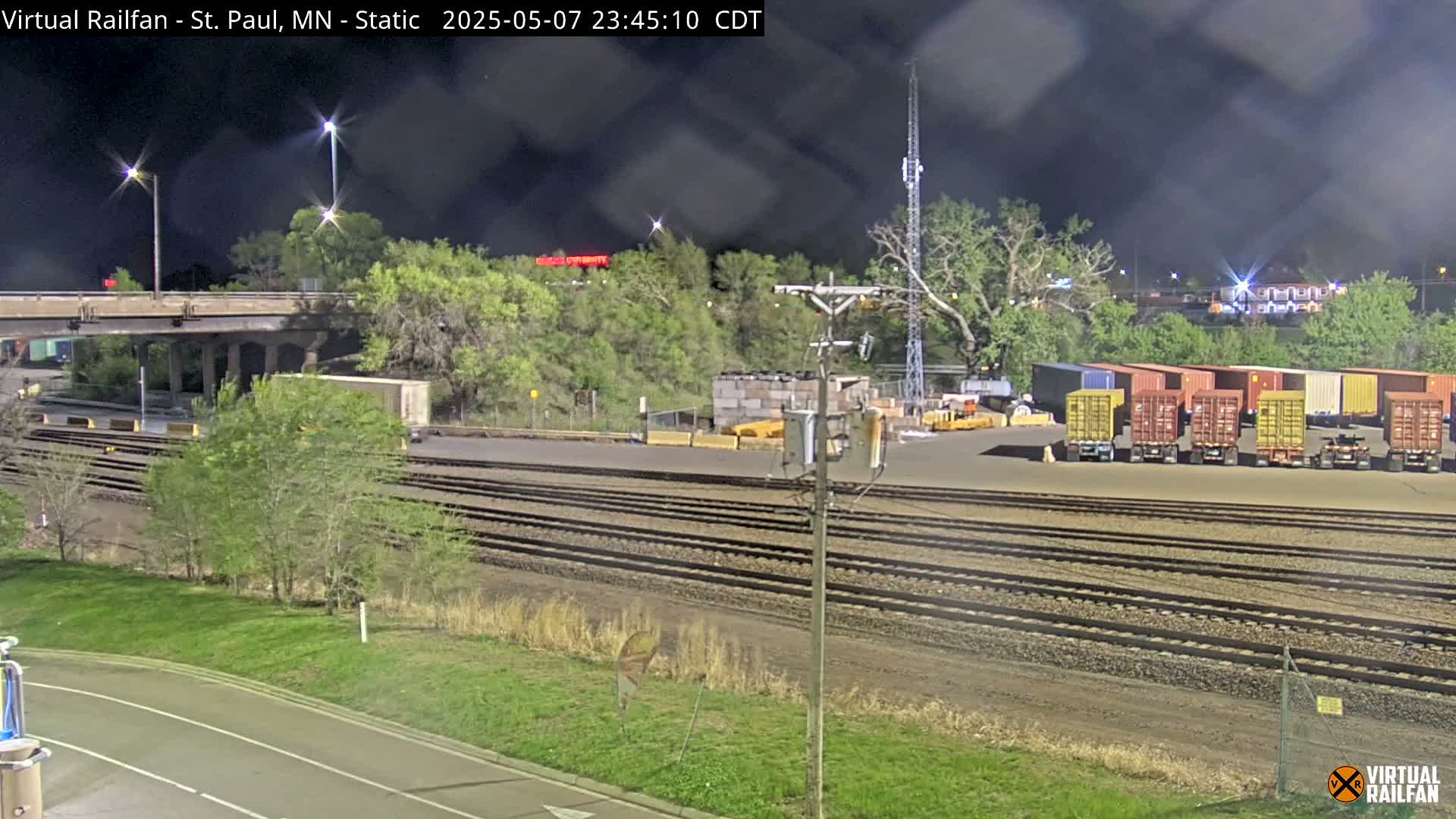 A nighttime view of railroad tracks and a freight yard with numerous shipping containers, under a partly cloudy sky.