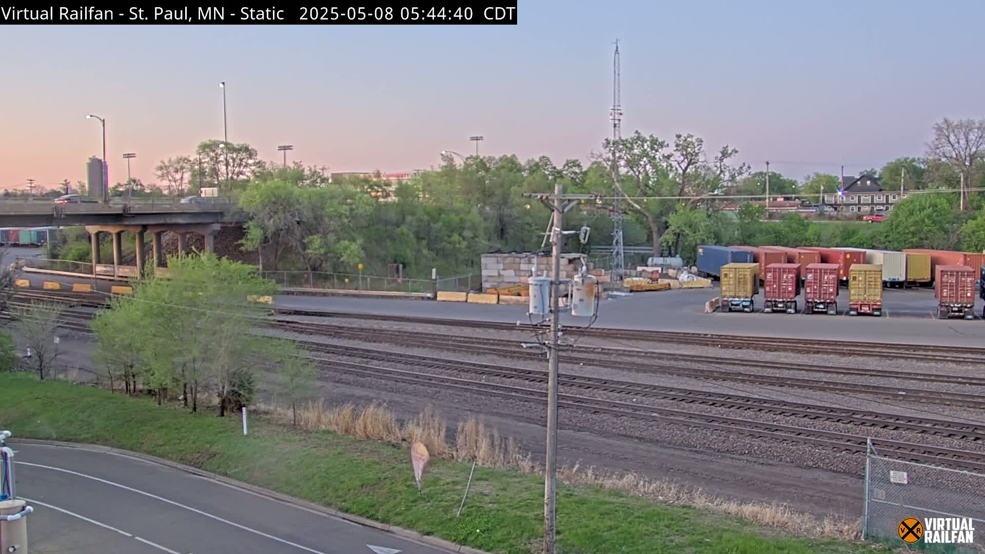 A twilight view of a rail yard with several shipping containers, train tracks, and a road under a bridge, surrounded by green trees and vegetation.