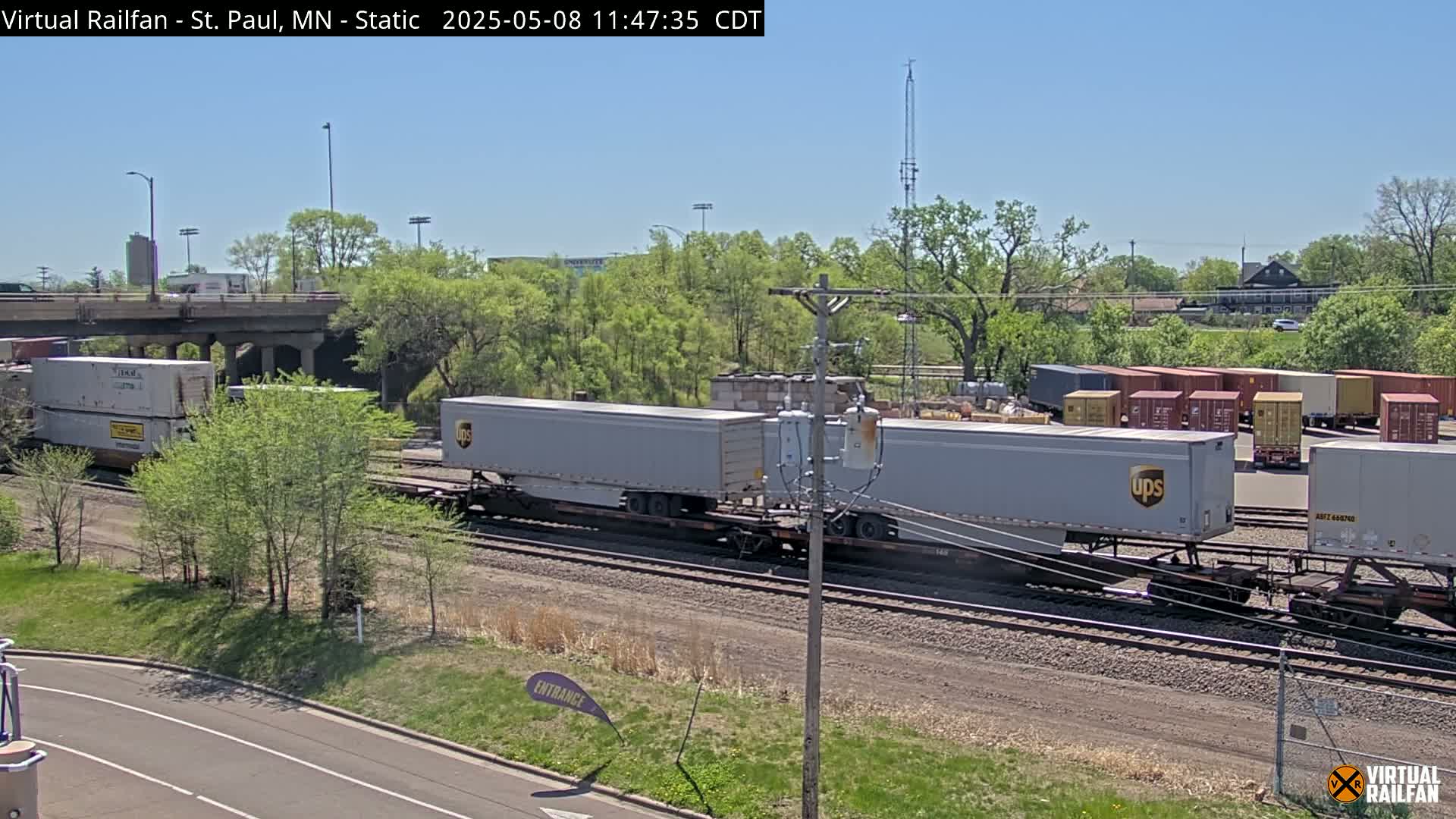 A freight train carrying UPS containers sits on railway tracks under a sunny sky next to a road and a line of trees.