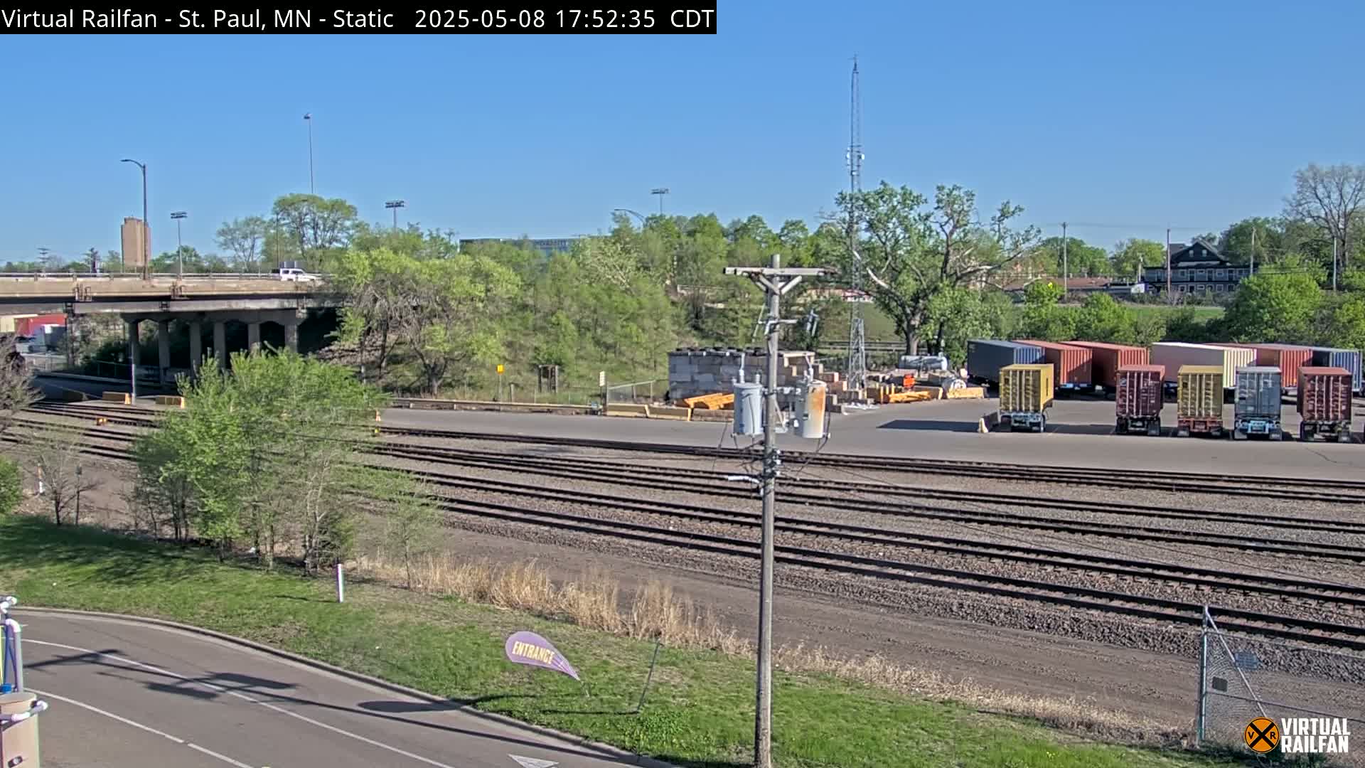Under a clear blue sky, a railway yard with multiple sets of train tracks is shown, alongside a road and a highway overpass, with several shipping containers parked in the yard and lush green trees in the background.