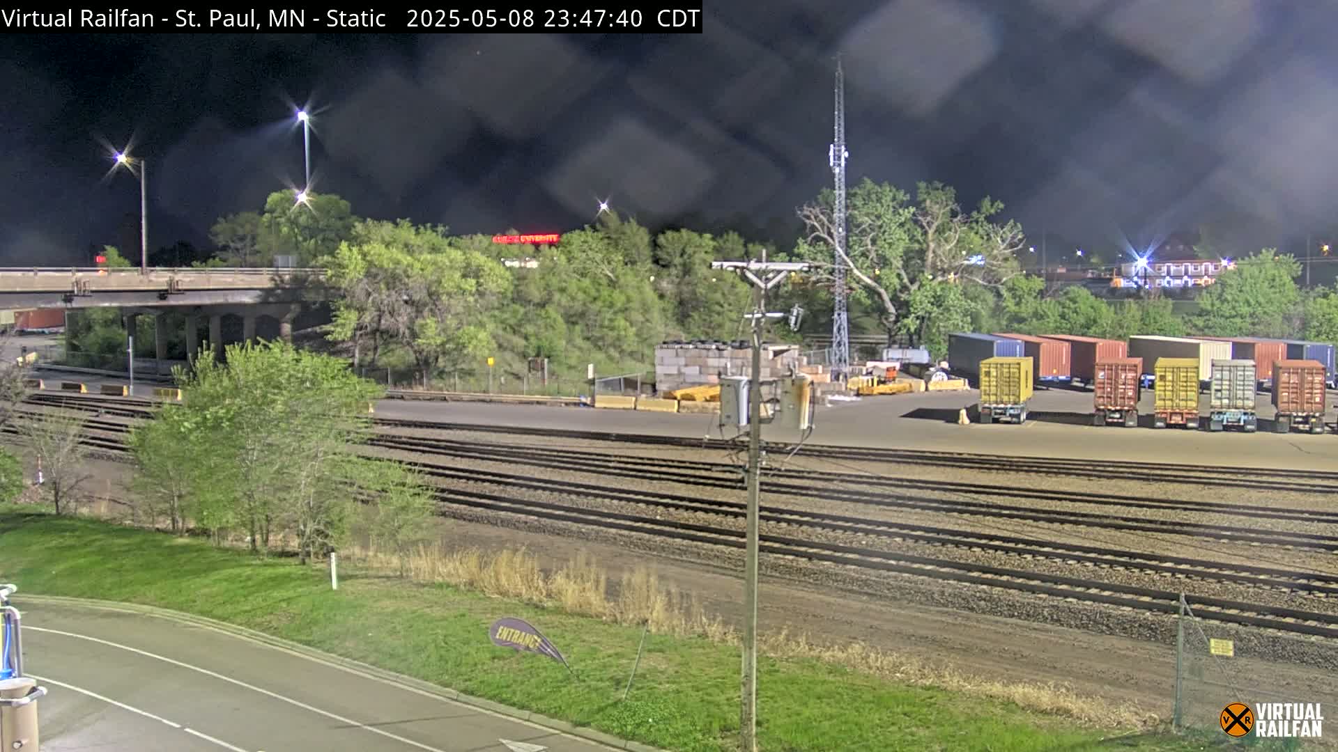 A nighttime view of a rail yard with several cargo containers, train tracks, and vegetation under a dark, cloudy sky.