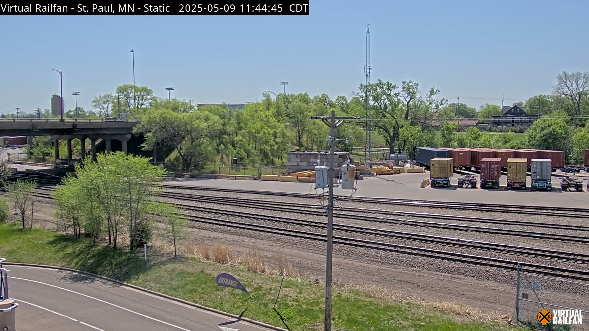 A sunny day shows a railroad yard with multiple train cars, a road, and a bridge over a wooded area.