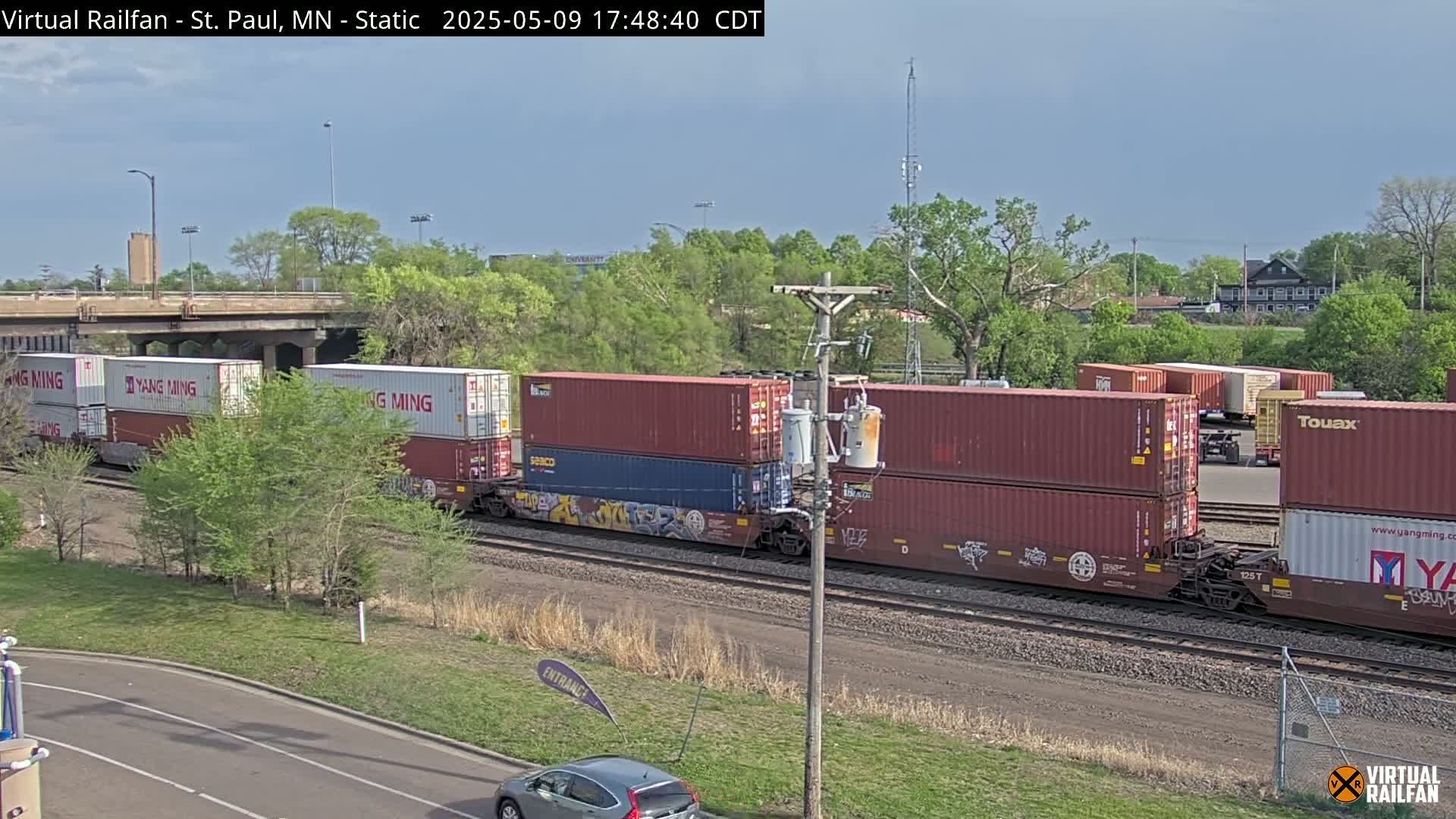 A freight train carrying stacked shipping containers moves along a railway track under a partly cloudy sky, next to a road and lush green vegetation.