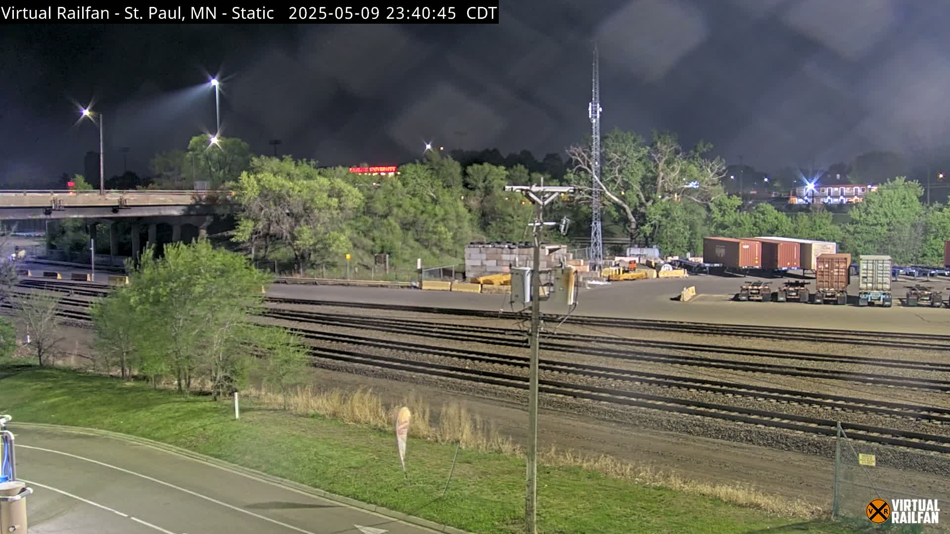 Under a dark, cloudy night sky, a nighttime view shows a rail yard with several shipping containers, train tracks, and a roadway overpass, surrounded by trees and vegetation.