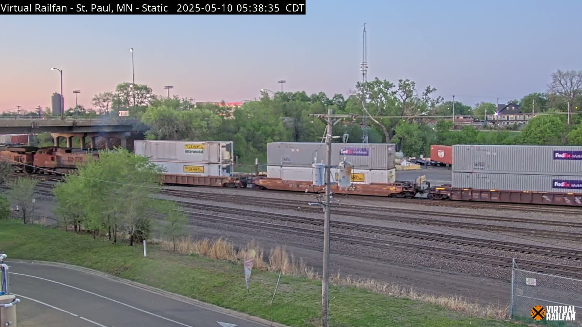 A freight train carrying several shipping containers is on railroad tracks next to a road under a highway overpass at dawn.
