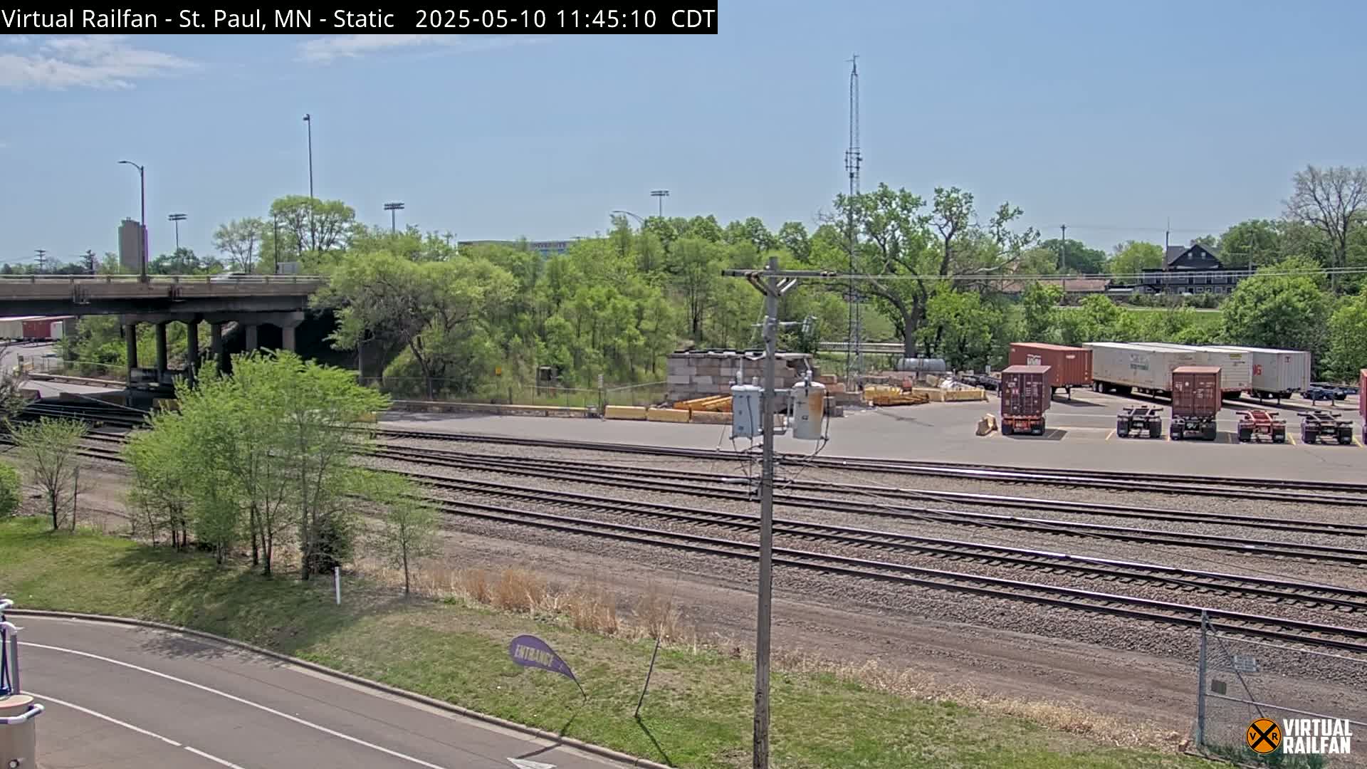 Under a clear blue sky, railroad tracks run alongside a road, passing under a bridge and near a yard where several cargo containers and trailers are parked next to lush green trees.