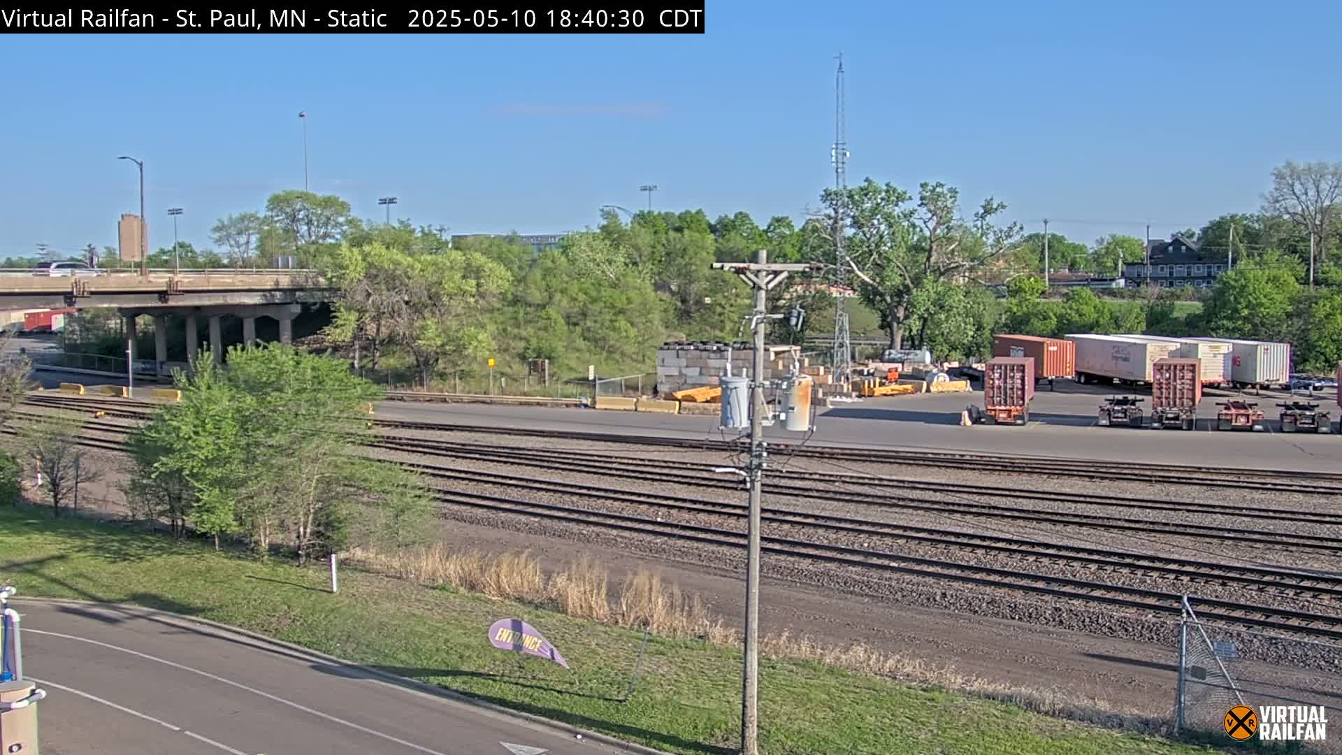 Under a clear blue sky, a view of railroad tracks and several shipping containers sits beside a road and a bridge, surrounded by green trees and vegetation.