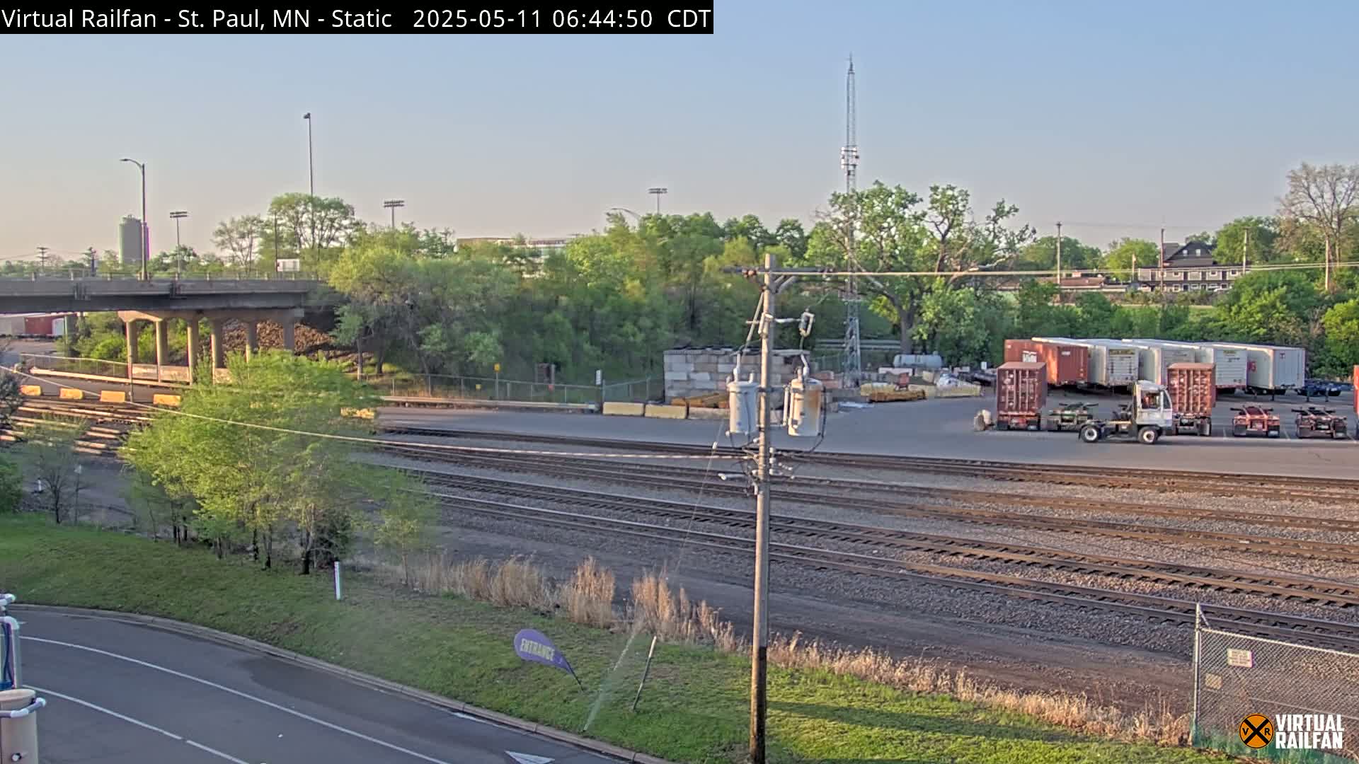 Under a clear, bright sky, a railway yard with several freight containers and a road beside multiple sets of train tracks are visible, with lush green trees and vegetation surrounding the area.