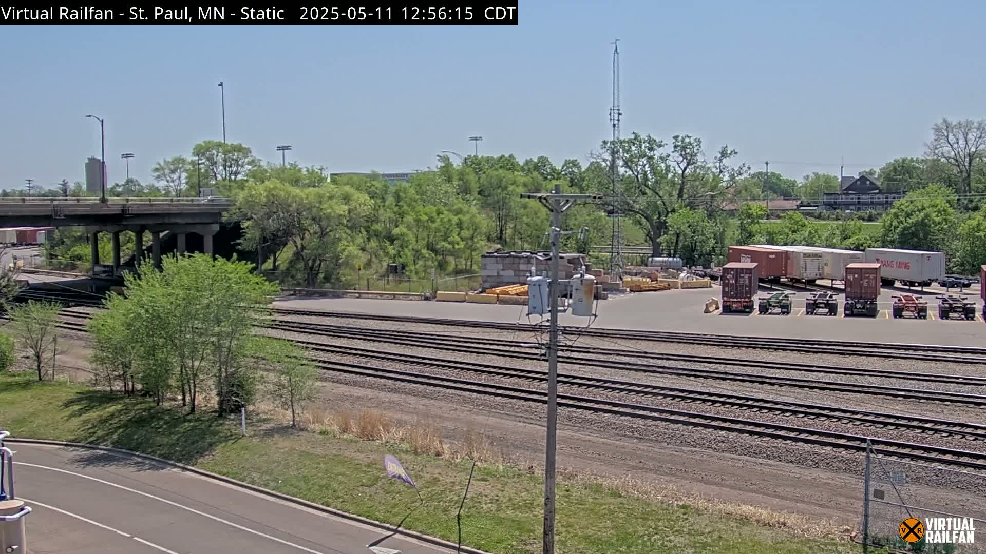 A sunny day reveals a railway yard with multiple train tracks, several shipping containers, and a road overpass, all surrounded by lush green trees.