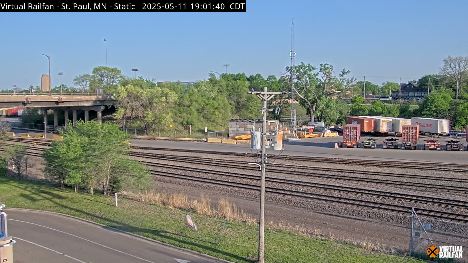 Under a clear sky, a view of railroad tracks, a roadway, and several shipping containers sits near a bridge and some trees.