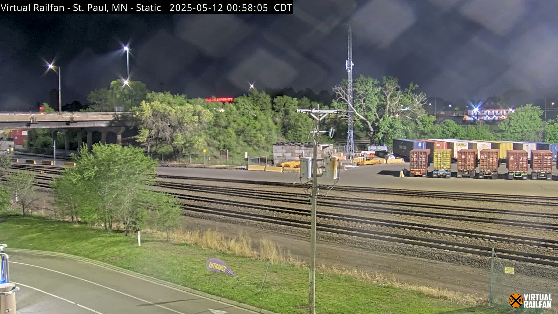 A nighttime view of a rail yard with multiple shipping containers, train tracks, and a highway overpass under a partly cloudy sky.