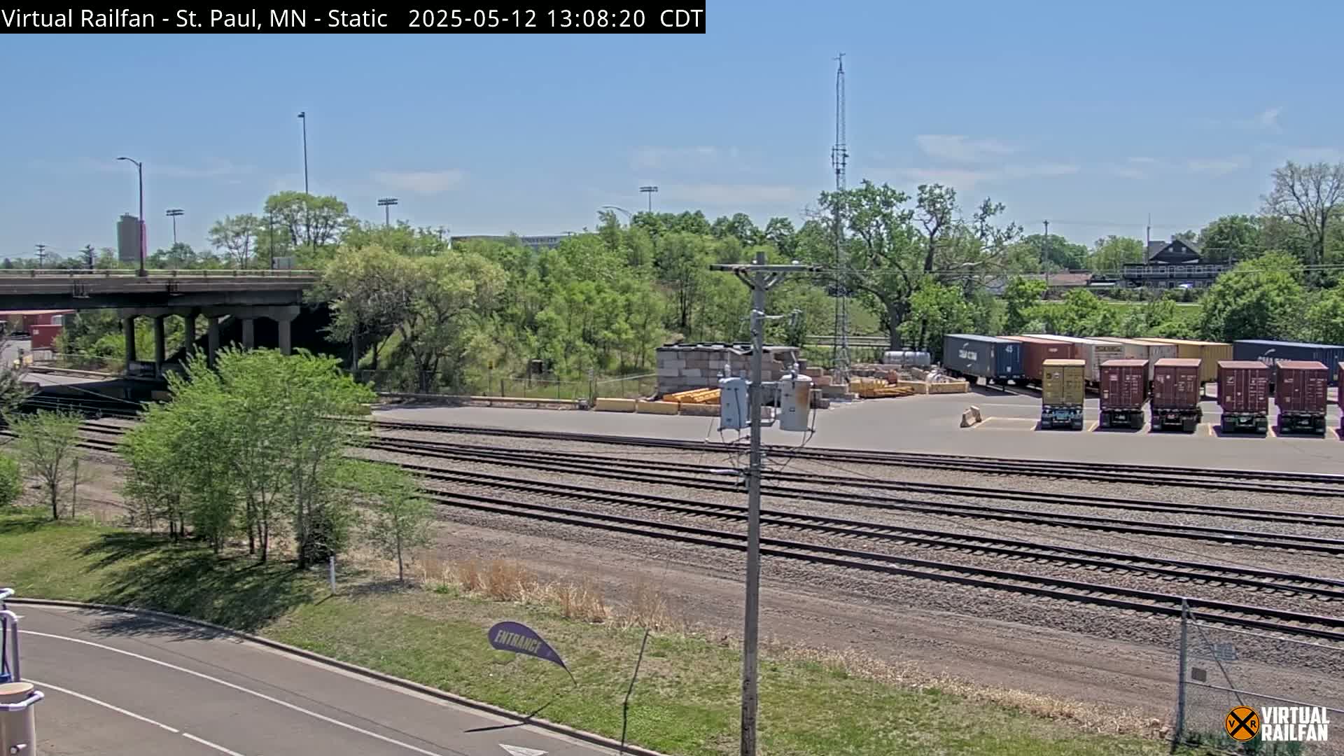 Under a clear blue sky, a railroad yard with multiple sets of train tracks is shown, alongside a road, a bridge, and several shipping containers parked nearby.
