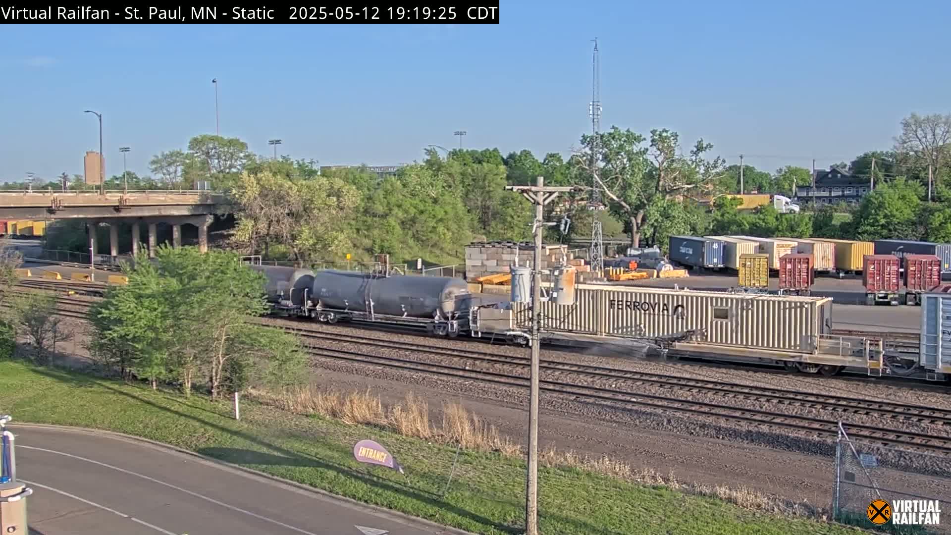 A freight train carrying tanker cars and shipping containers is traveling along a railway under a highway overpass on a sunny day.