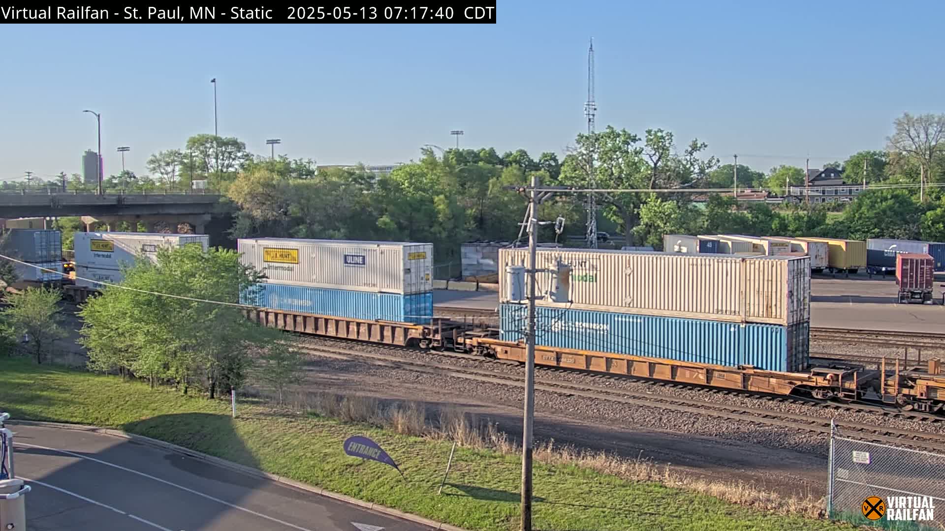Two trains carrying stacks of shipping containers are on railroad tracks under a clear, sunny sky, surrounded by green trees and a roadway.