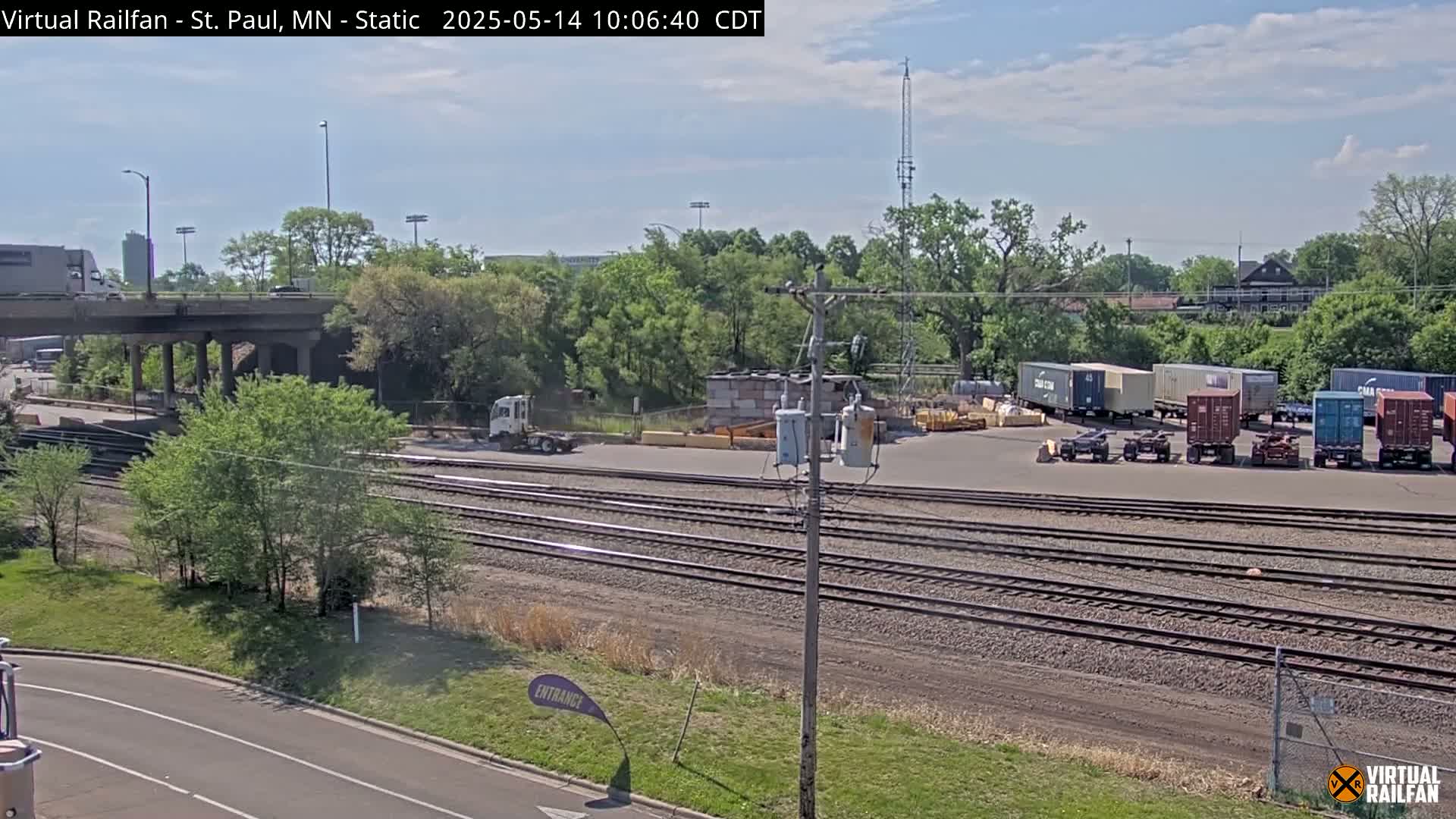 Under a partly cloudy sky, a view of railroad tracks, a road, several cargo containers, and a highway overpass with some trees and vegetation surrounding the area.
