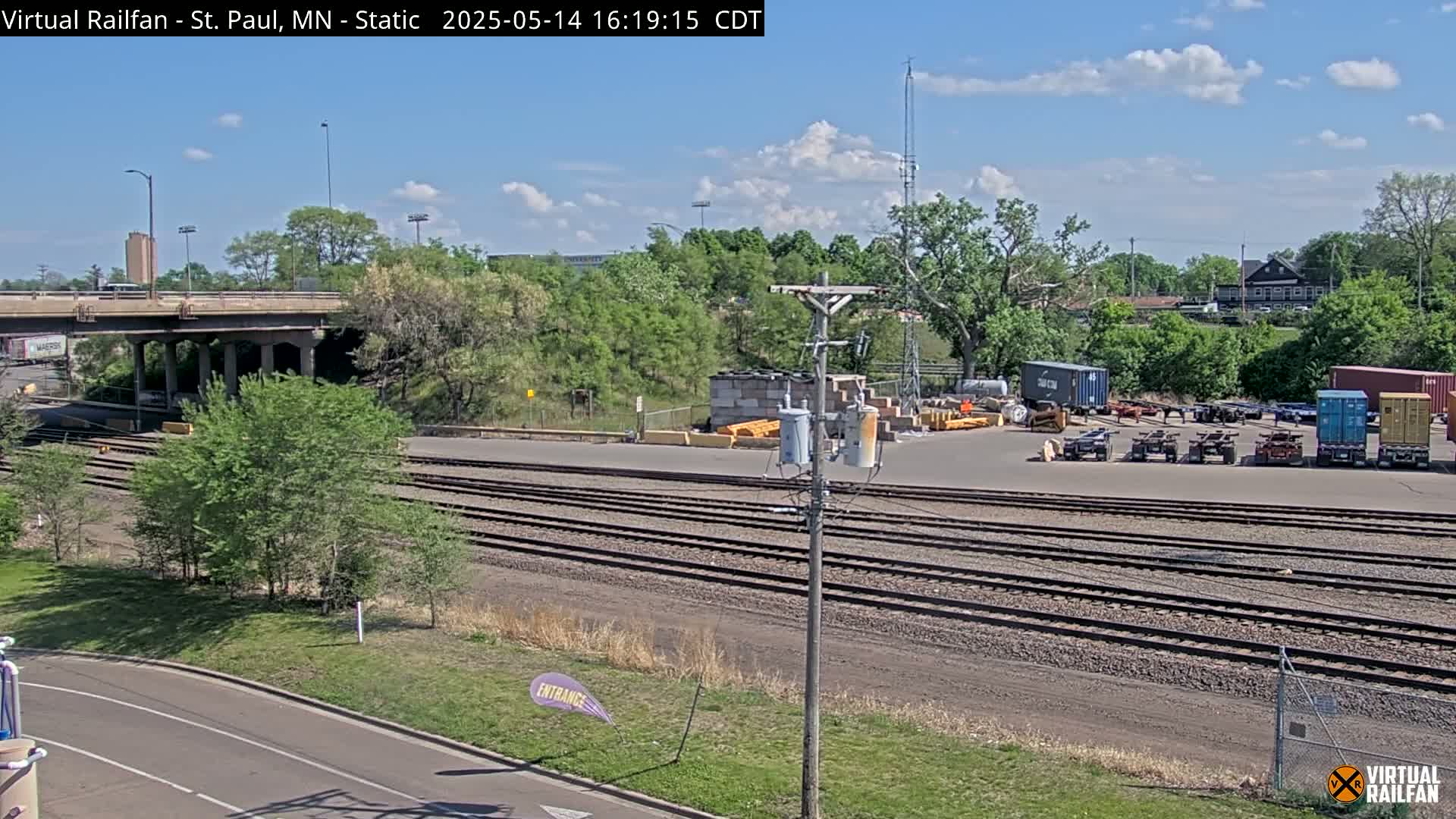 A sunny day reveals a railroad yard with multiple sets of train tracks, a road, and several shipping containers and trailers near a treed area and overpass.