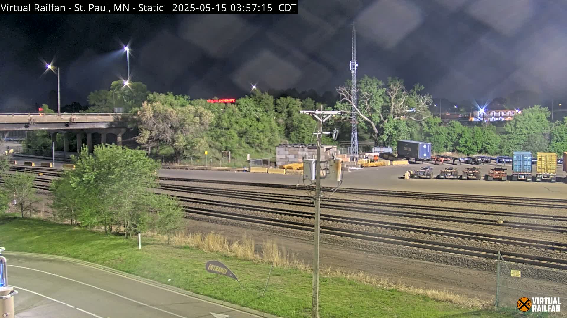 A nighttime view of a railway yard with several shipping containers and train tracks under a partly cloudy sky.