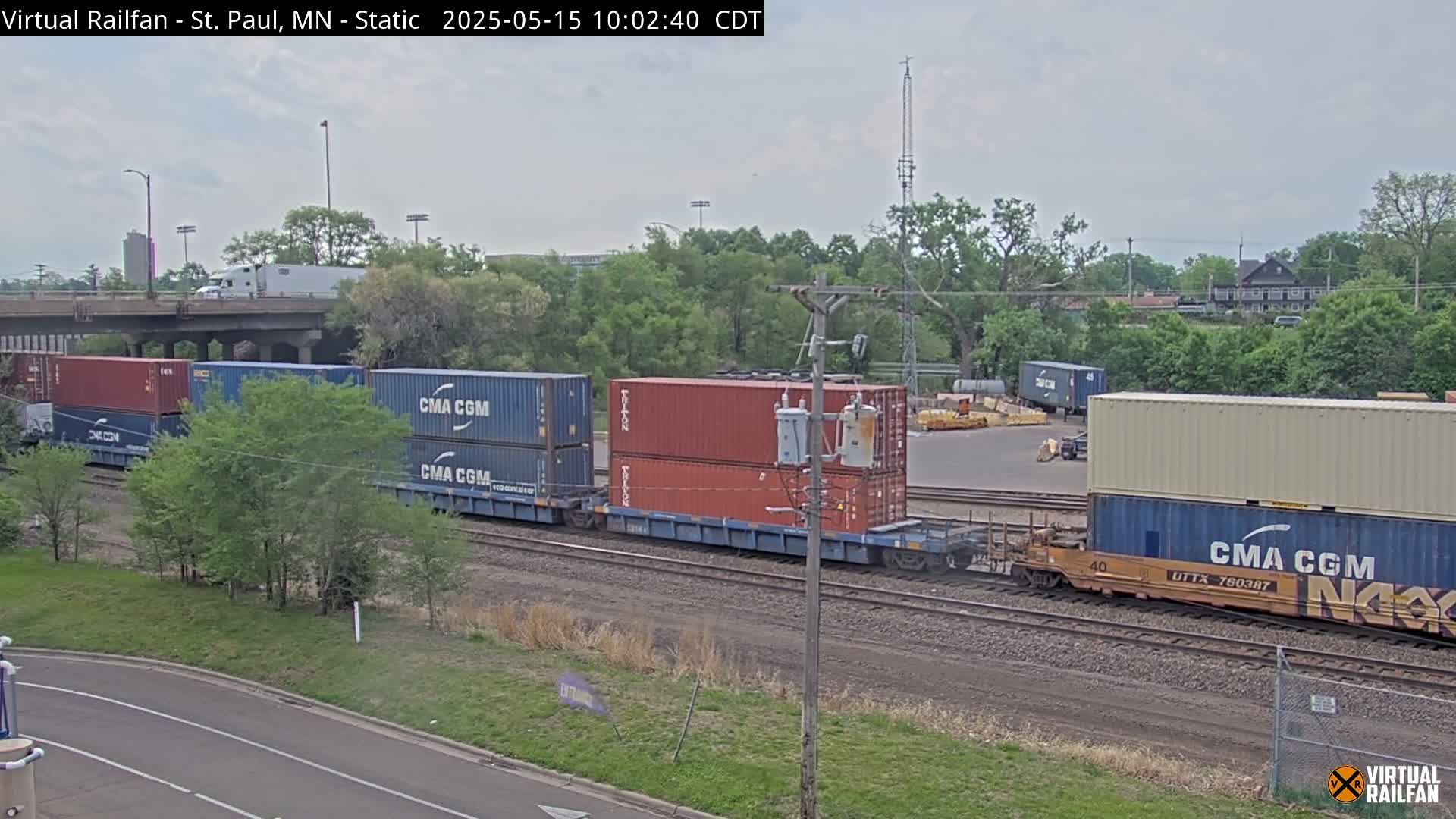 A freight train carrying multiple shipping containers sits on railroad tracks under a partly cloudy sky, next to a road and lush green trees.