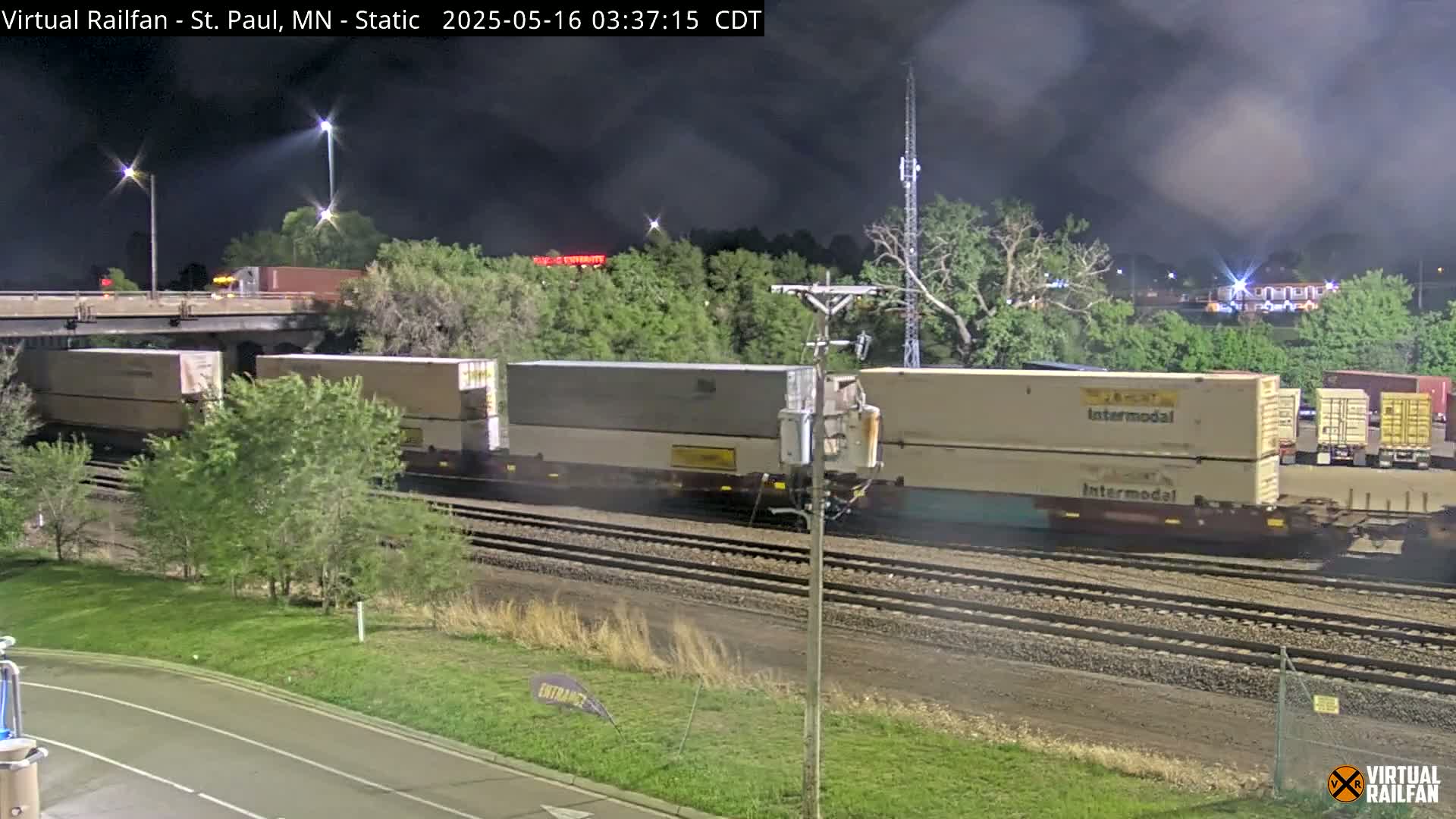 A freight train carrying several intermodal containers moves along a track at night under a cloudy sky.