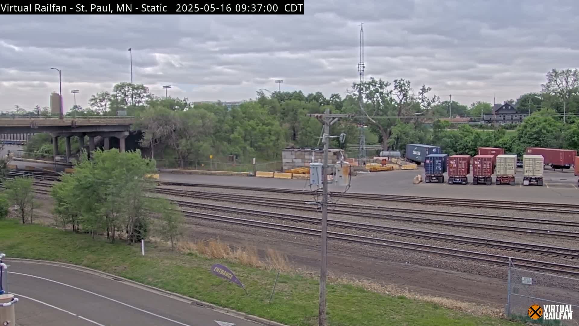 A cloudy day shows railroad tracks, a road, a bridge, several shipping containers, and lush green trees.