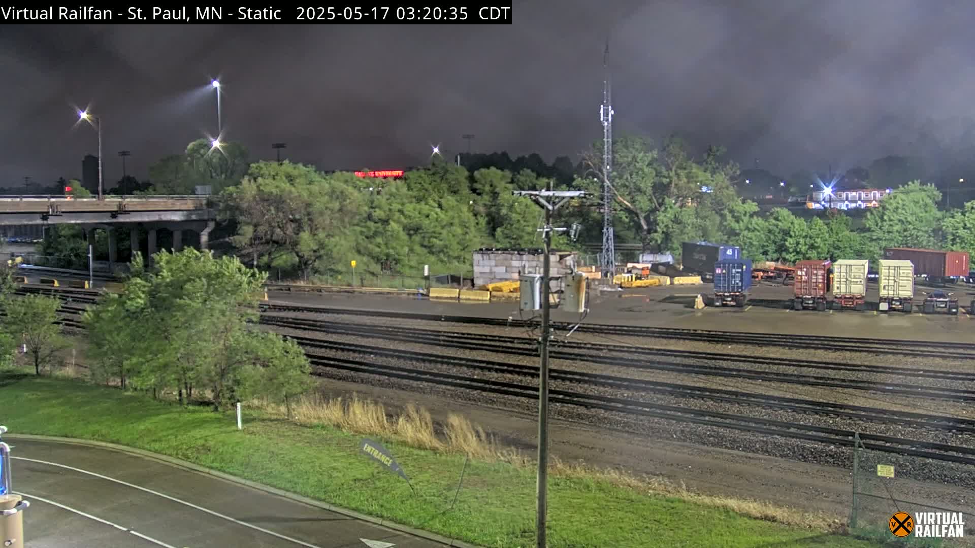 A nighttime view of a rail yard with several shipping containers, under a dark, cloudy sky.