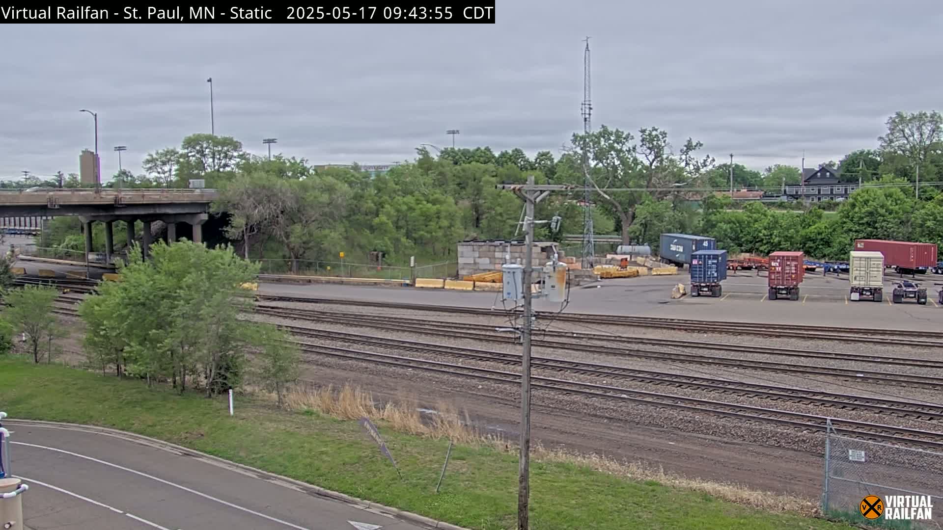 A railroad yard with several shipping containers and multiple sets of train tracks is visible under an overcast sky.