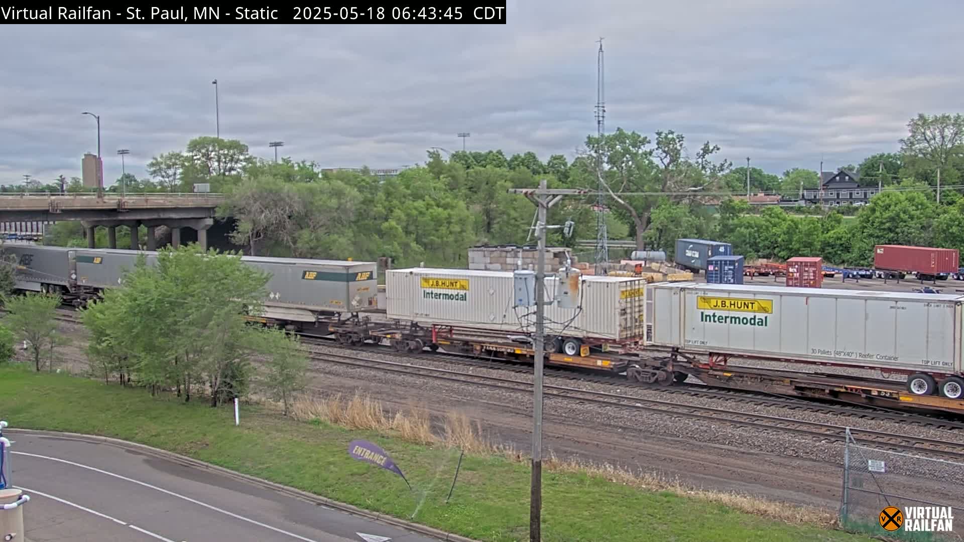 A freight train carrying several large shipping containers moves along railroad tracks under an overcast sky, surrounded by lush green trees and a bridge.