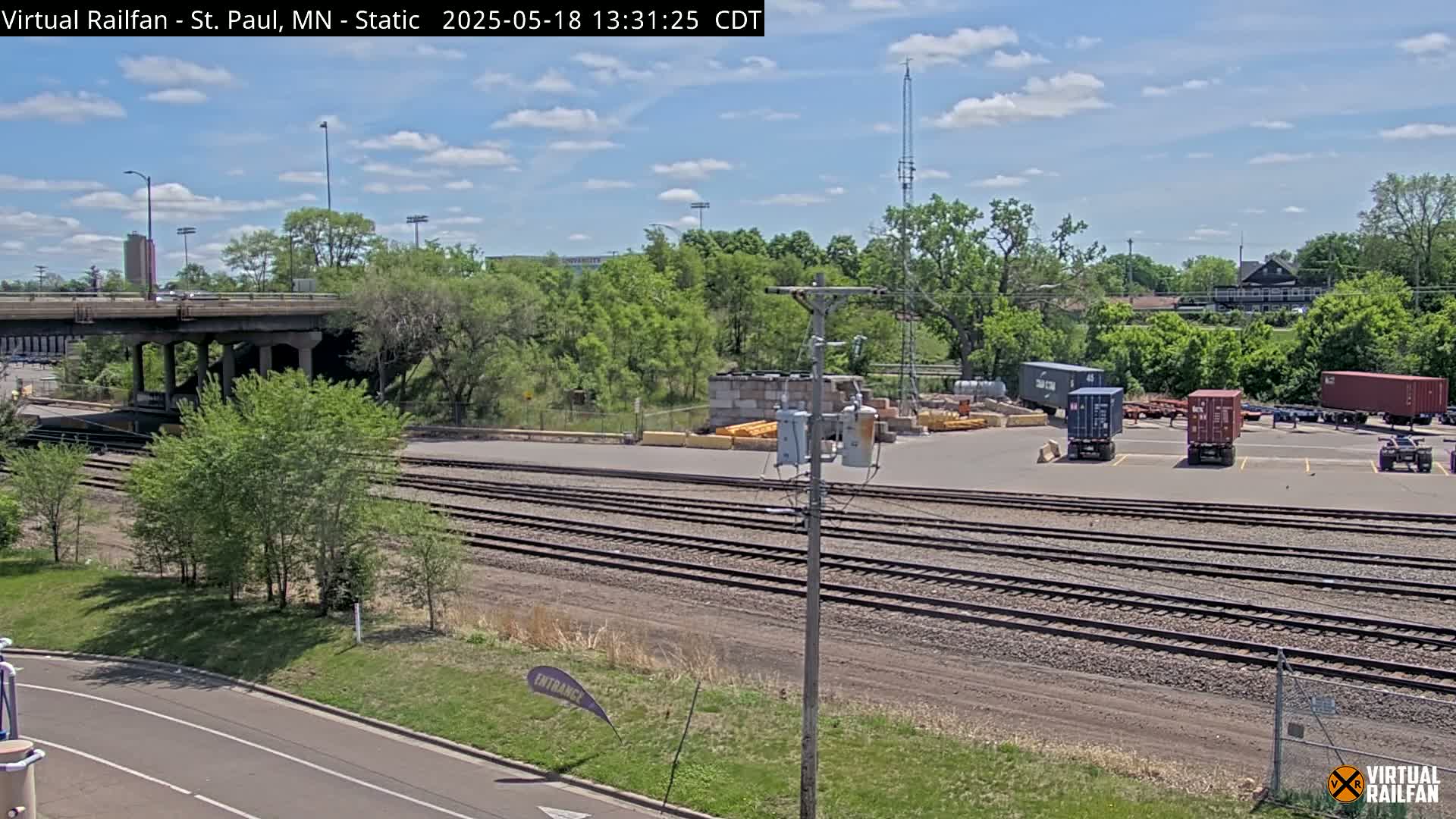 A sunny day shows a railroad yard with several shipping containers, train tracks, a bridge, and lush green trees under a partly cloudy sky.