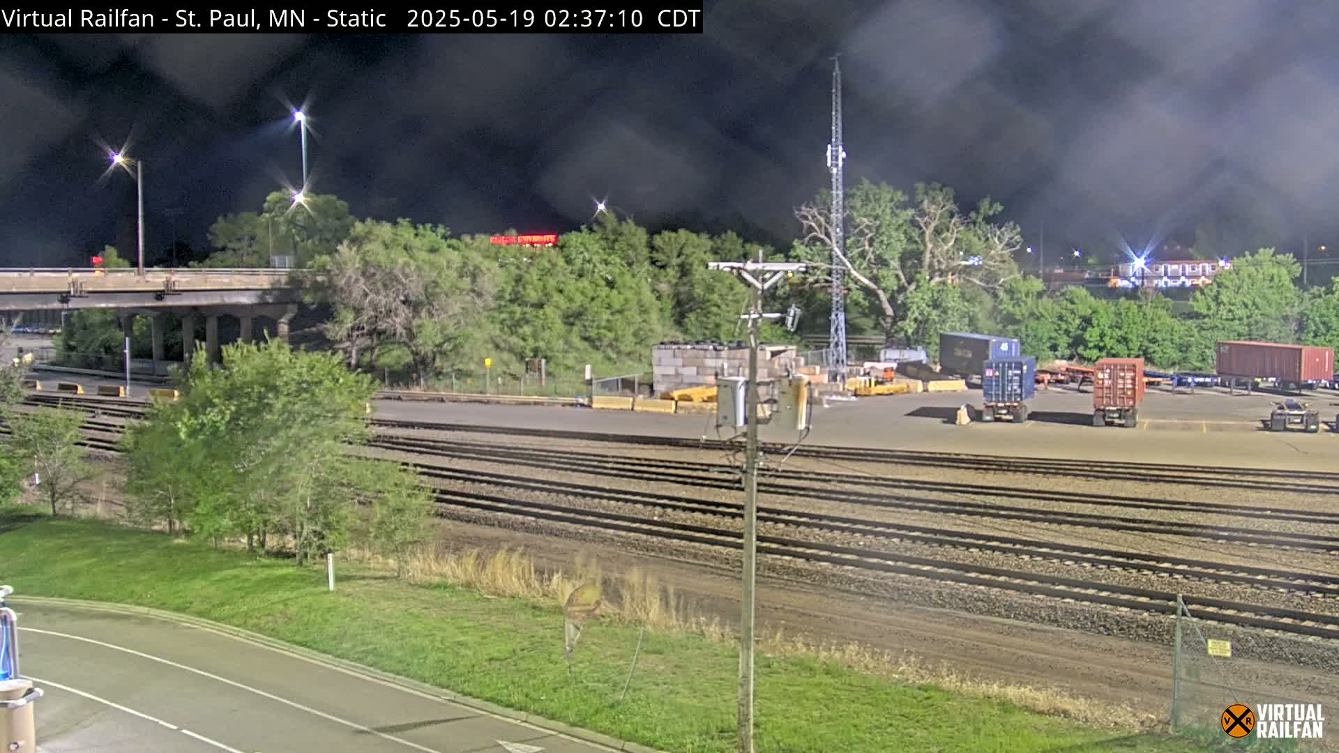 A nighttime view of a train yard with multiple sets of railroad tracks, several shipping containers, and a highway overpass in the background under a dark, cloudy sky.