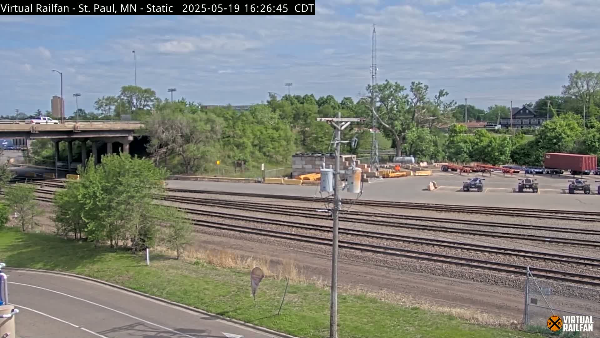 A partly cloudy day reveals a rail yard with multiple sets of train tracks, a road, and a bridge overhead, surrounded by green trees and some parked vehicles.