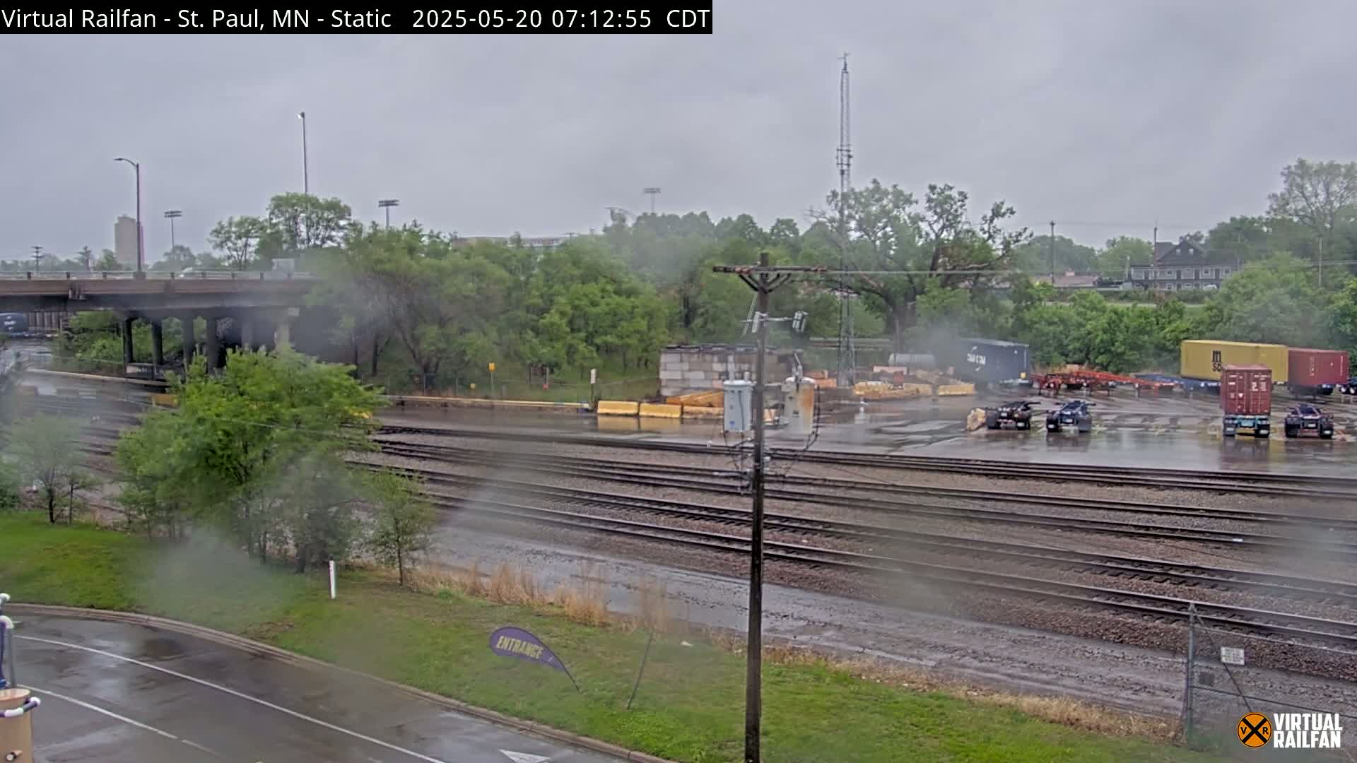 A rainy scene shows multiple sets of train tracks, a road, and several vehicles parked near some buildings and greenery, all under a gray sky.