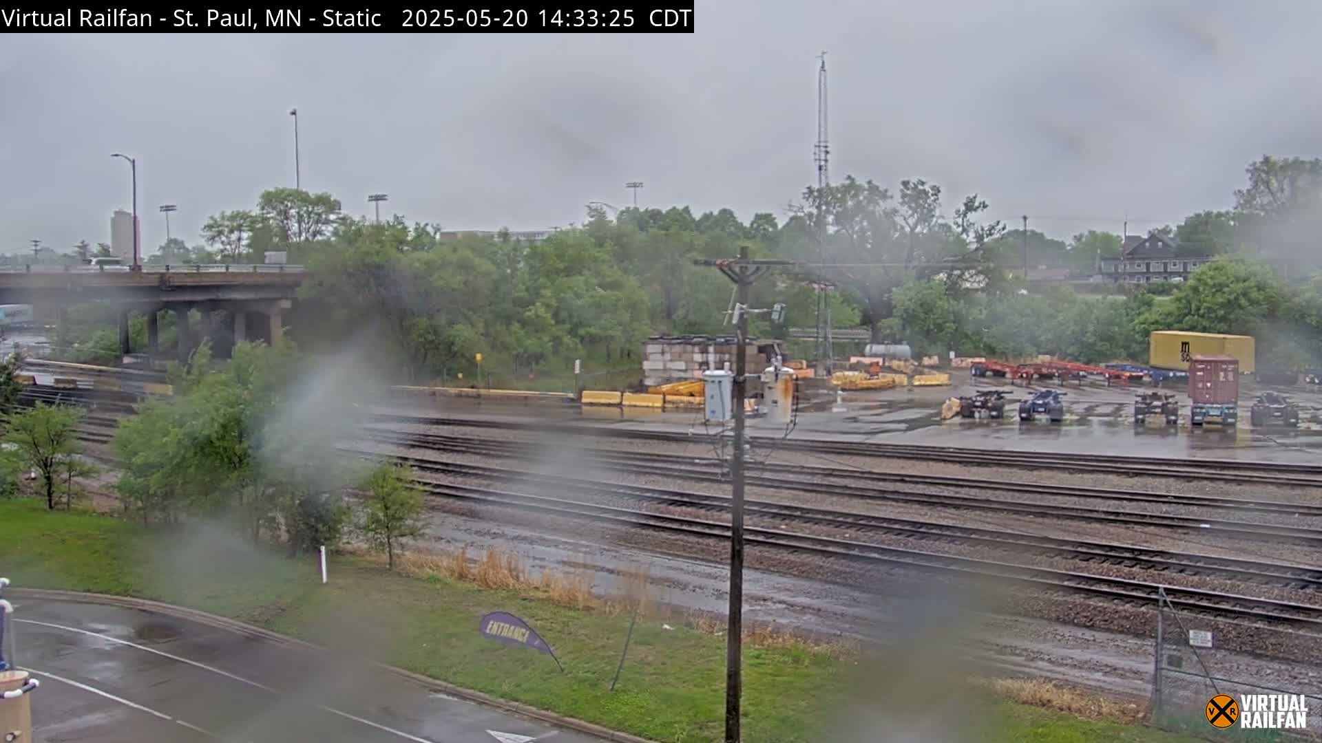 A rainy view of railroad tracks, a road, and a yard with several semi-trailers and other equipment.