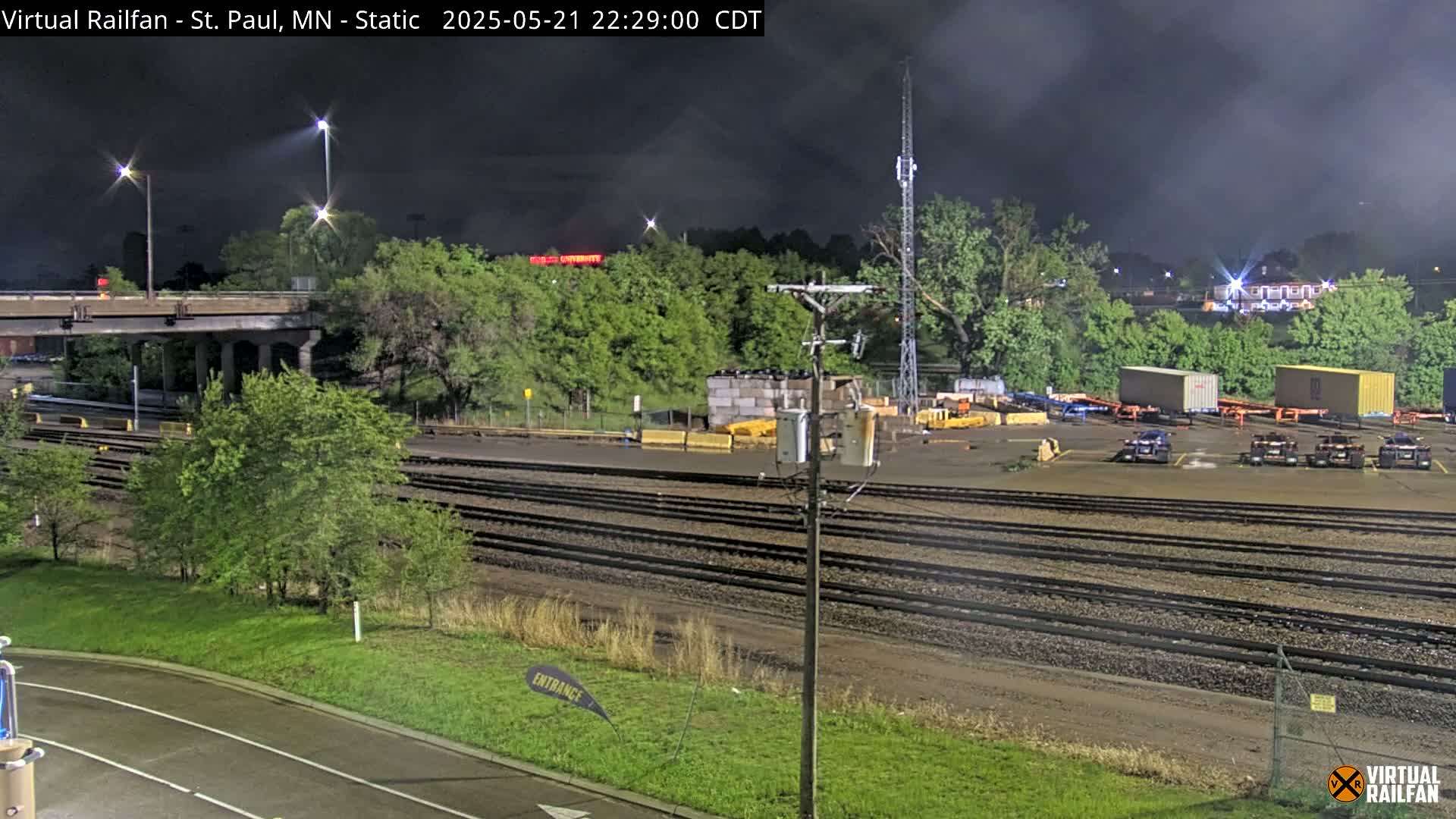 A nighttime view of a railroad yard with multiple sets of tracks, some containers, and green trees under a cloudy sky.