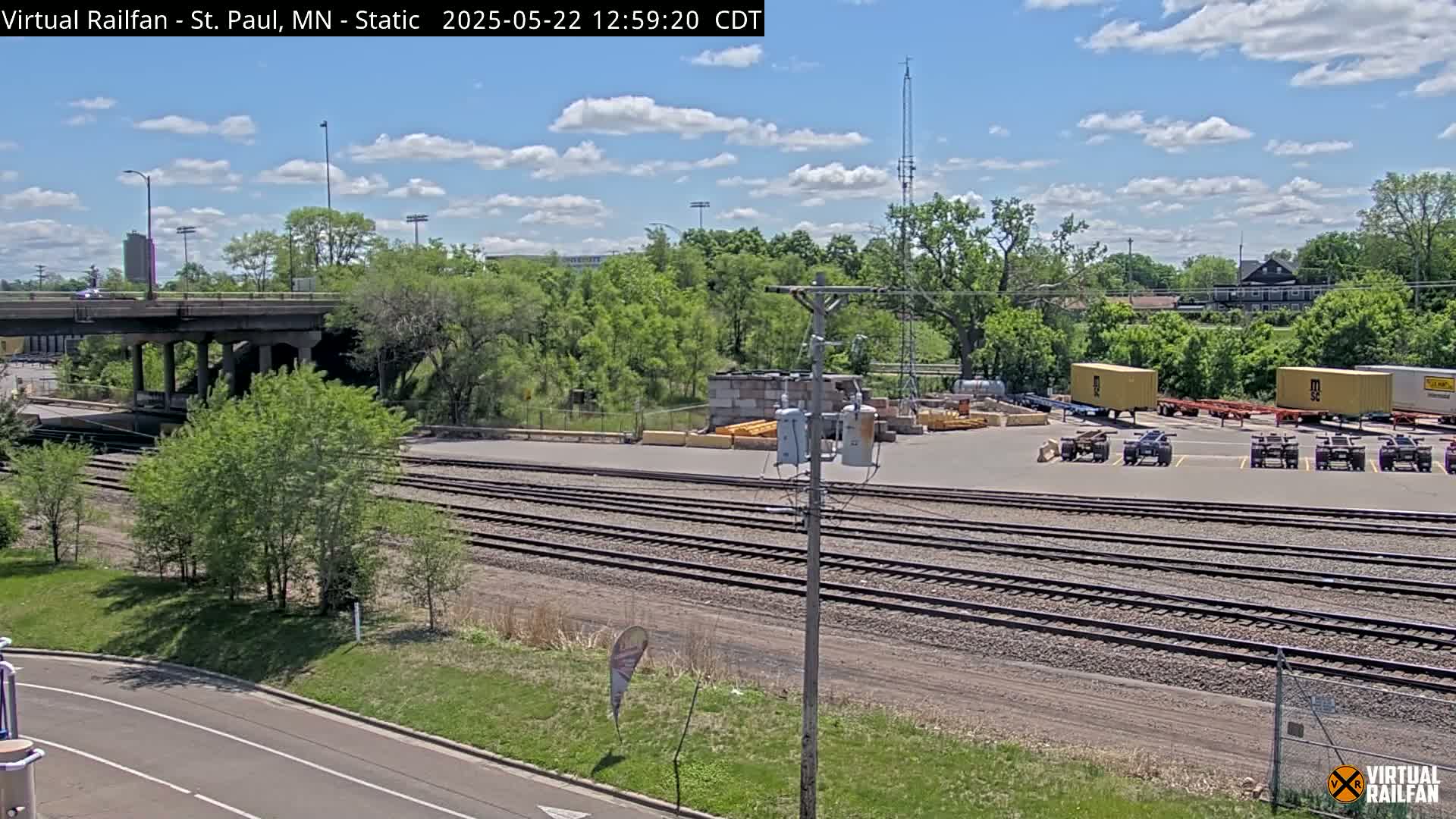A sunny day shows a view of railroad tracks, a road, a bridge, and several shipping containers parked in a lot, surrounded by green trees.