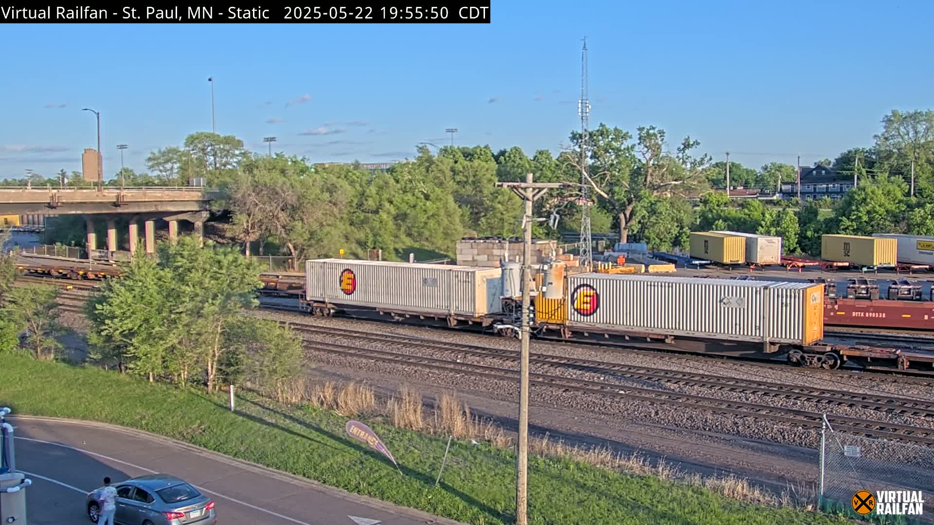 A long freight train carrying several large white shipping containers is passing under a highway overpass on a sunny day.