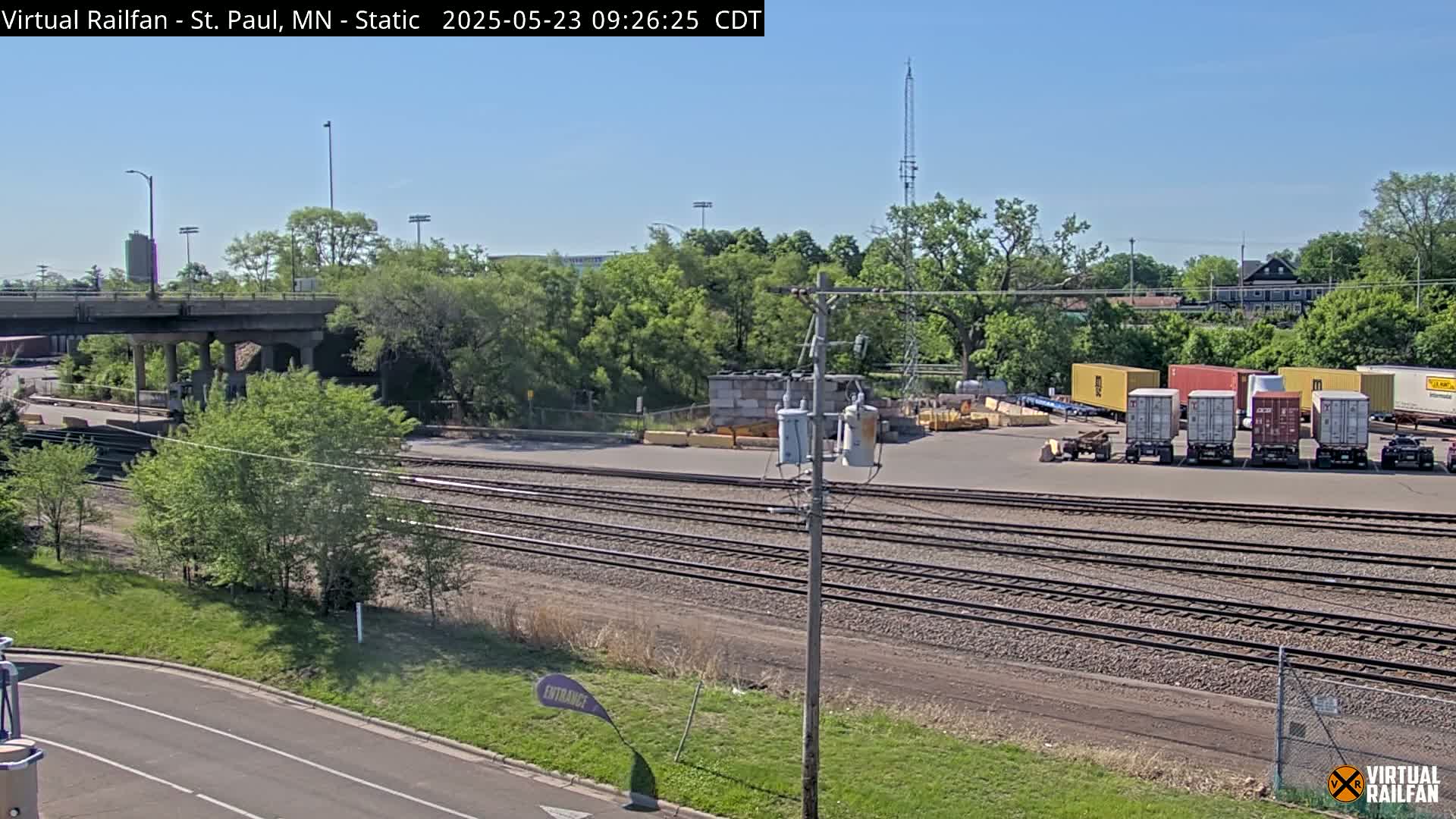 A sunny view of railroad tracks, a road, and several shipping containers near a tree-lined area under a clear sky.