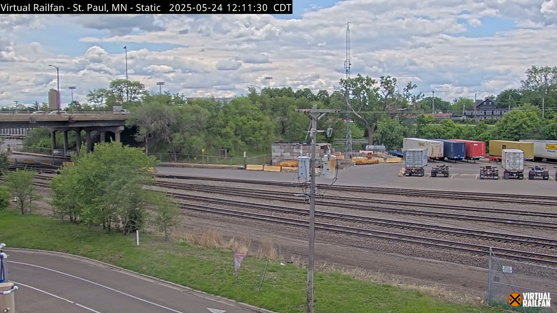 A partly cloudy day reveals a railway yard with several shipping containers, a road overpass, and lush green vegetation.