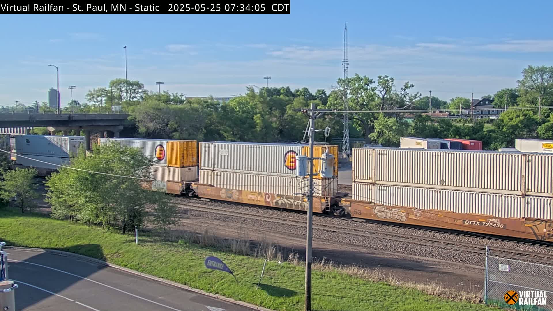 A freight train carrying multiple shipping containers moves along railroad tracks under a sunny sky, with green trees and a highway overpass in the background.