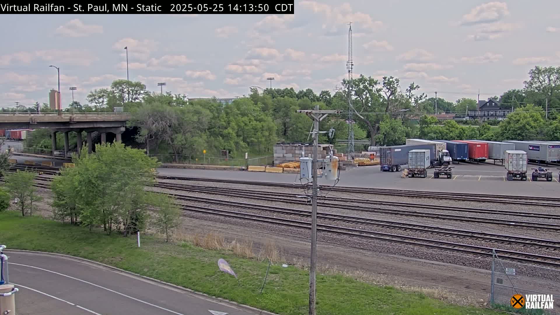 A partly cloudy day reveals a railway yard with multiple train tracks, a road, a bridge, and several shipping containers parked near a building surrounded by lush green trees.