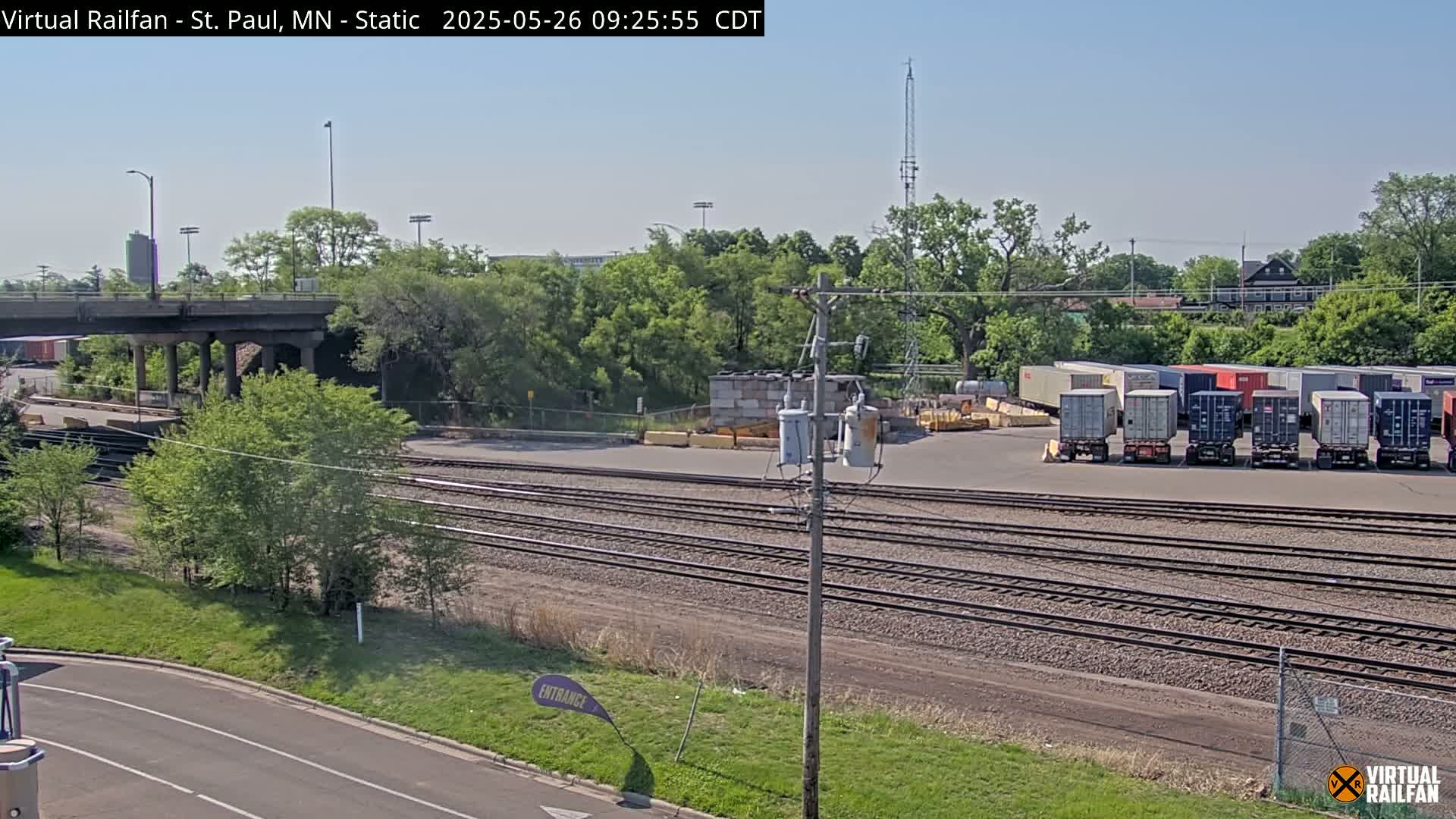 A sunny day reveals a railroad yard with multiple shipping containers, train tracks, and lush green vegetation surrounding a bridge and road.