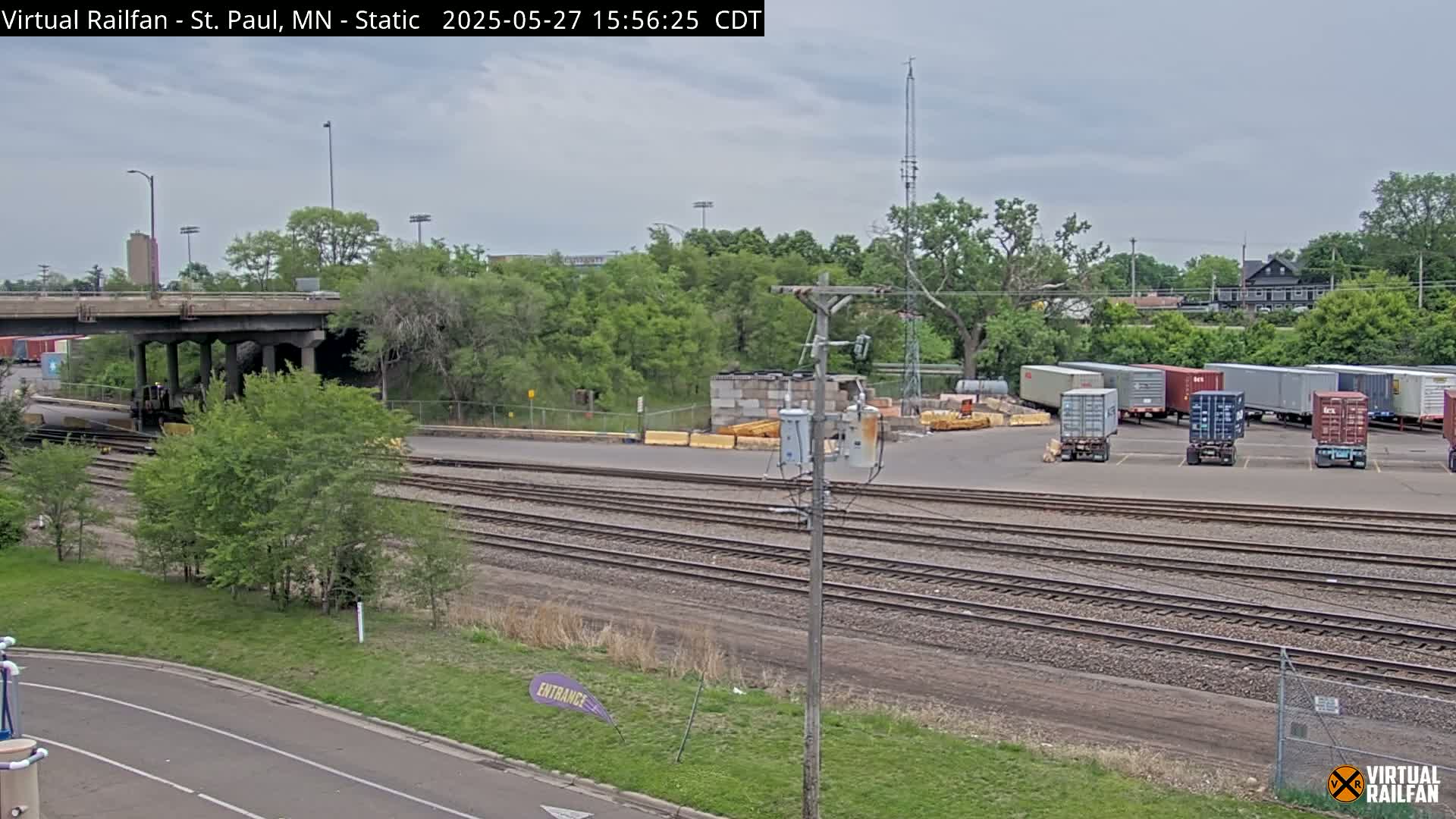 Under an overcast sky, a view of railroad tracks stretches toward a parking lot filled with shipping containers, adjacent to a bridge and lush green vegetation.